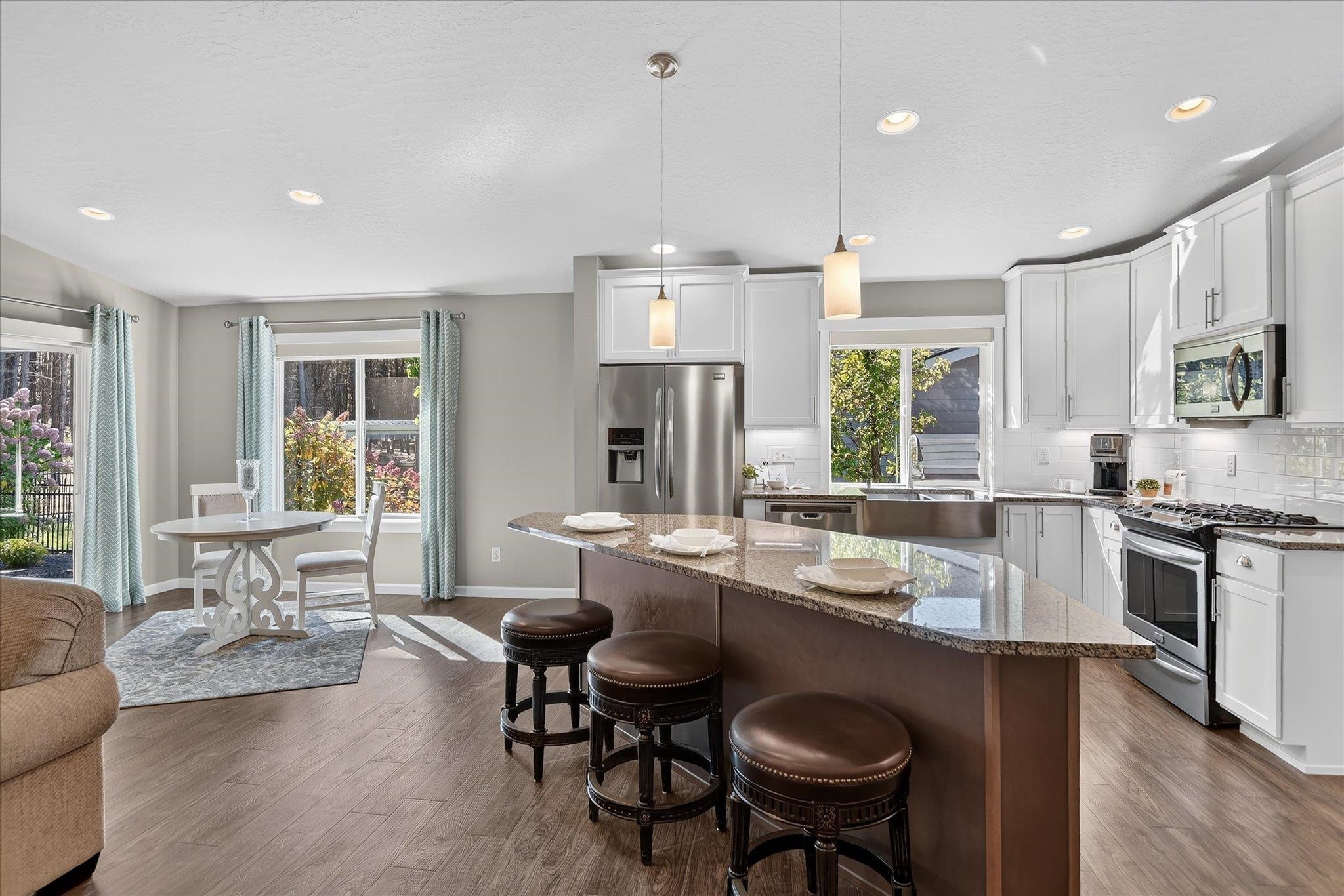 Bright kitchen with a granite island, three stools, white cabinetry, stainless appliances, and adjacent dining nook.