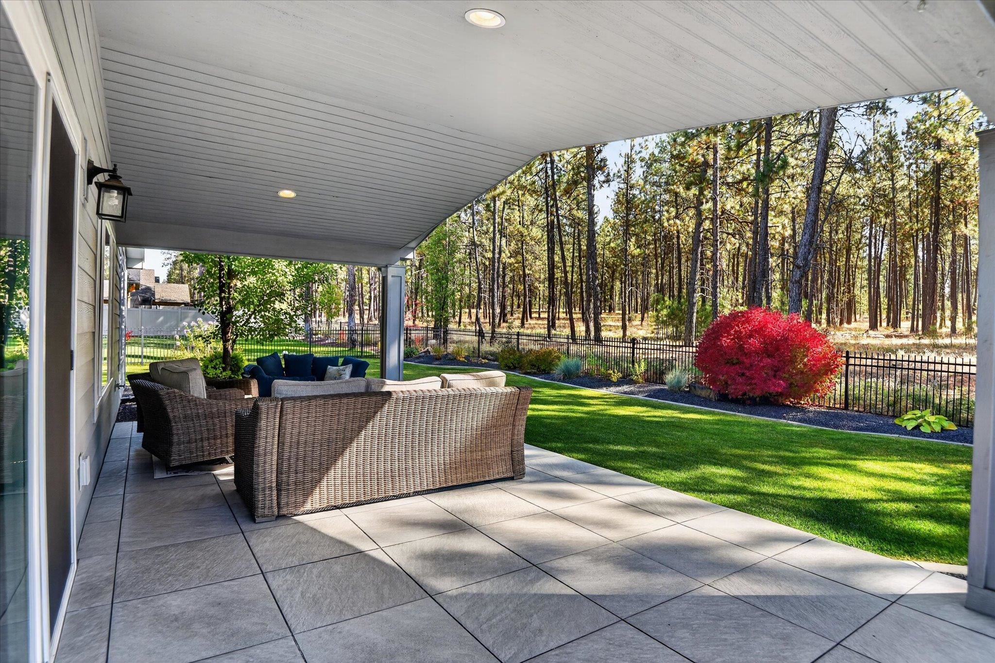 Covered patio with wicker seating overlooks a fenced lawn and pine trees, with a bright red shrub.