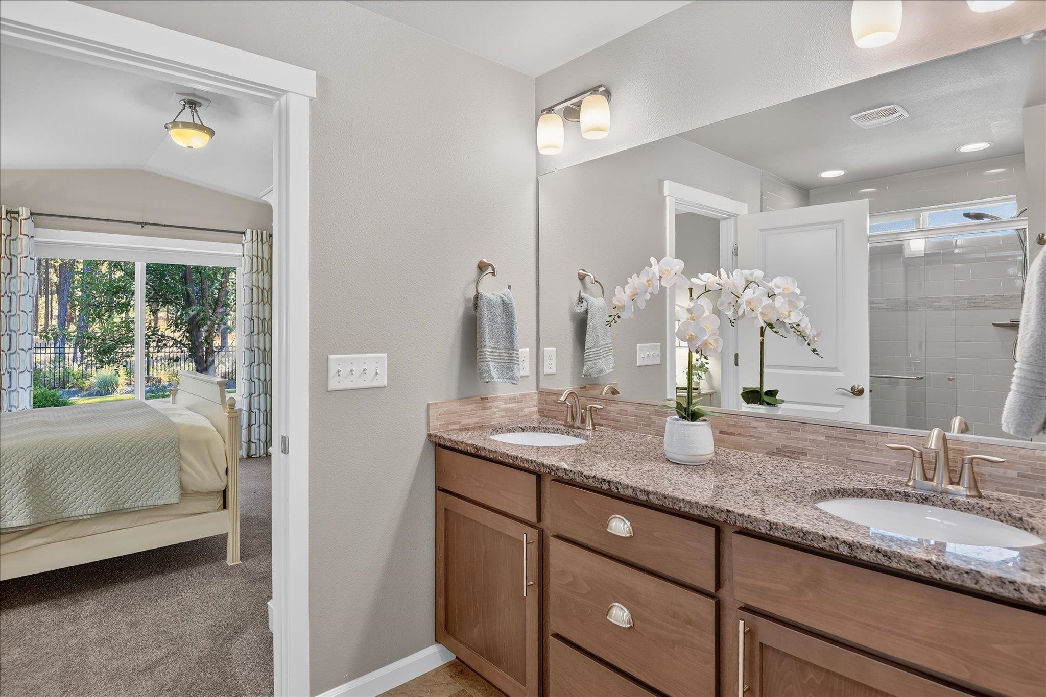 Bathroom with a double-sink granite vanity, tiled shower, and doorway to a bedroom.