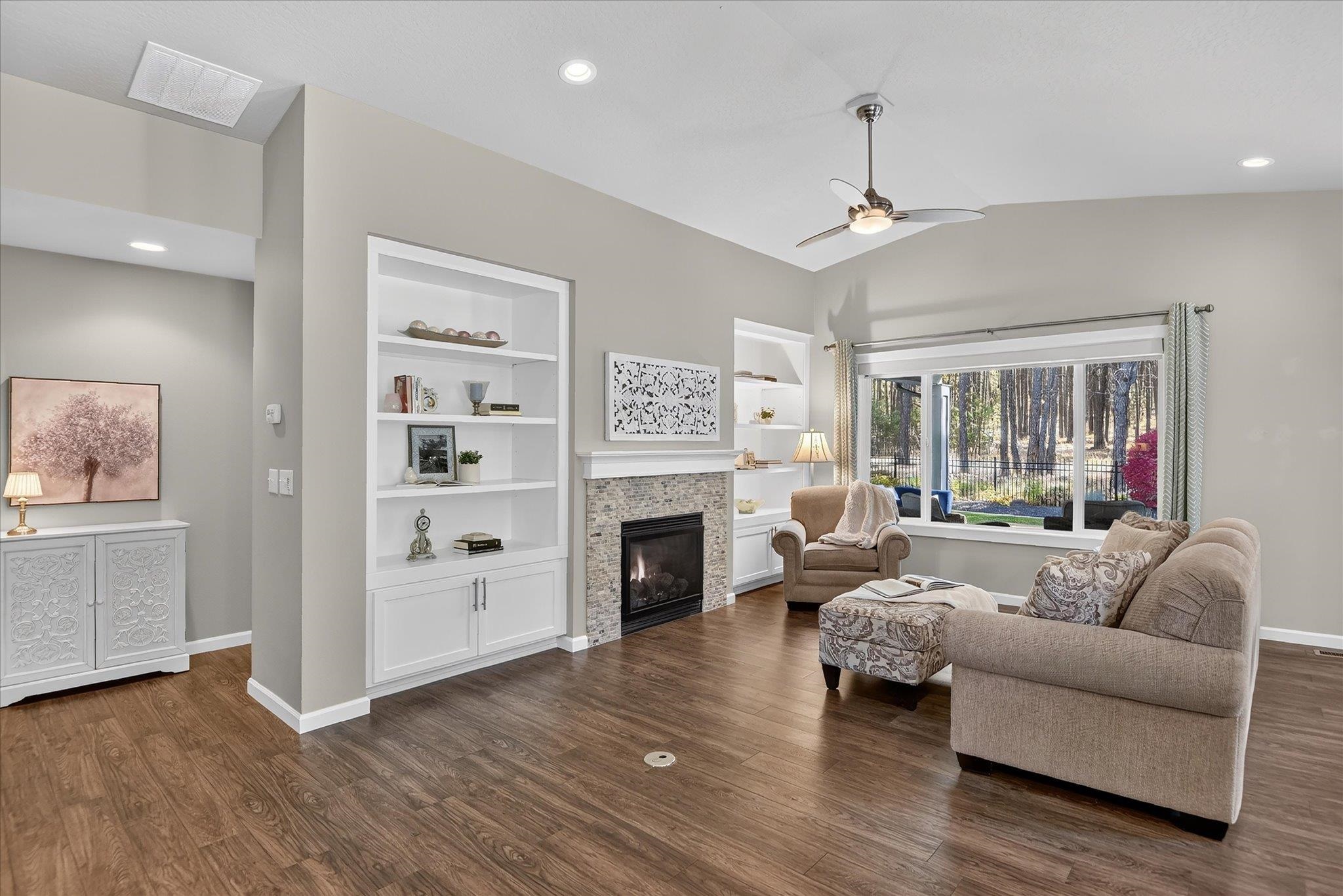 Living room with a stone fireplace, built-in shelves, hardwood floors, and a large window overlooking trees.
