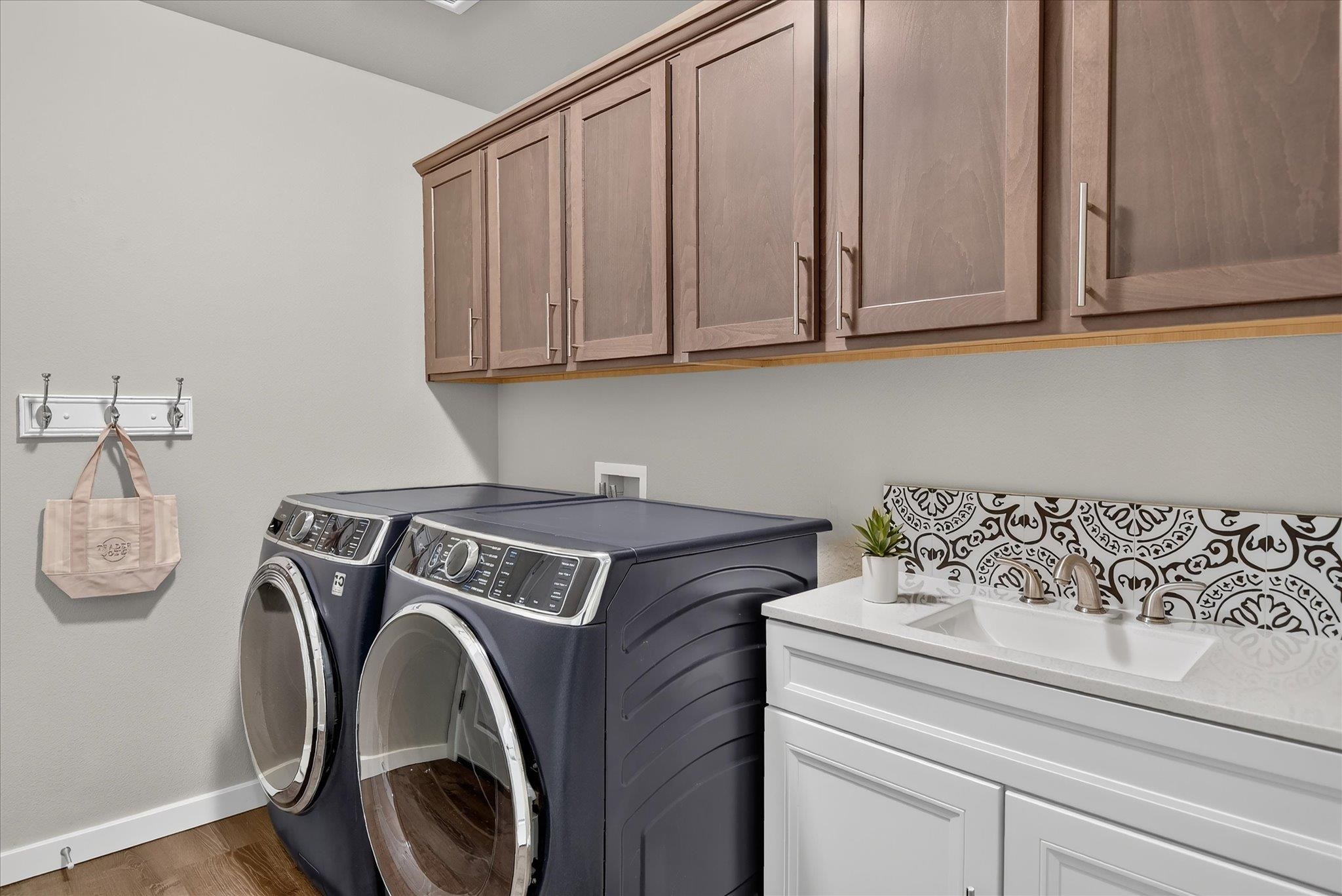 Laundry room with dark front-loading washer and dryer, wood cabinets, and a sink with patterned tile backsplash.