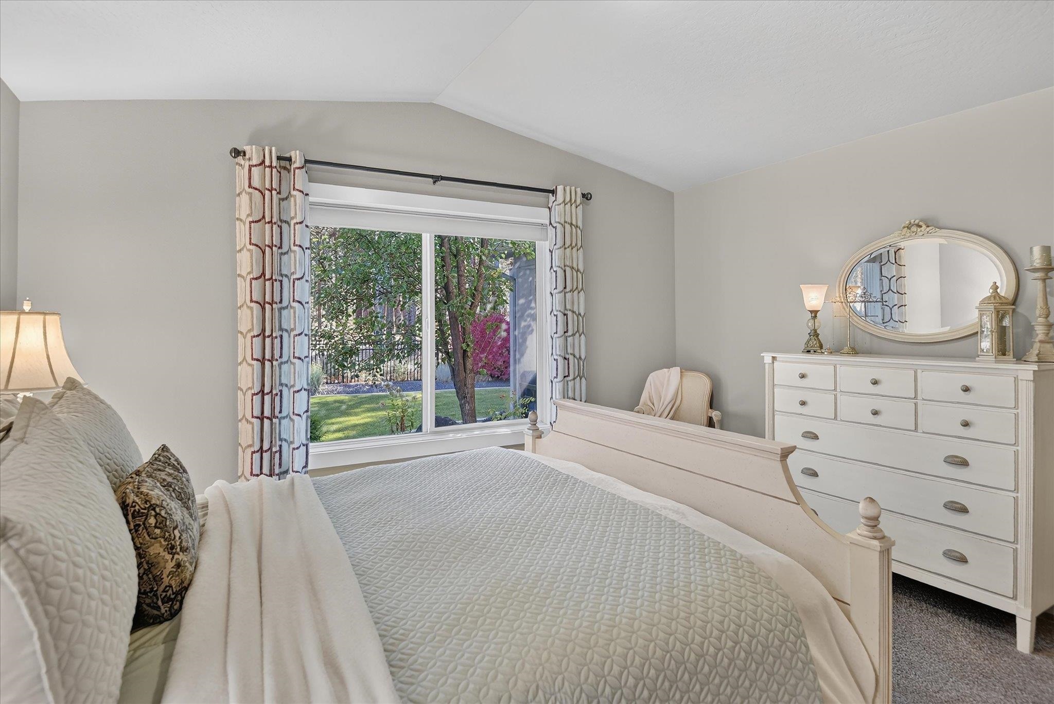 Bedroom with a white quilted bed, patterned curtains, and a large window overlooking a leafy yard.