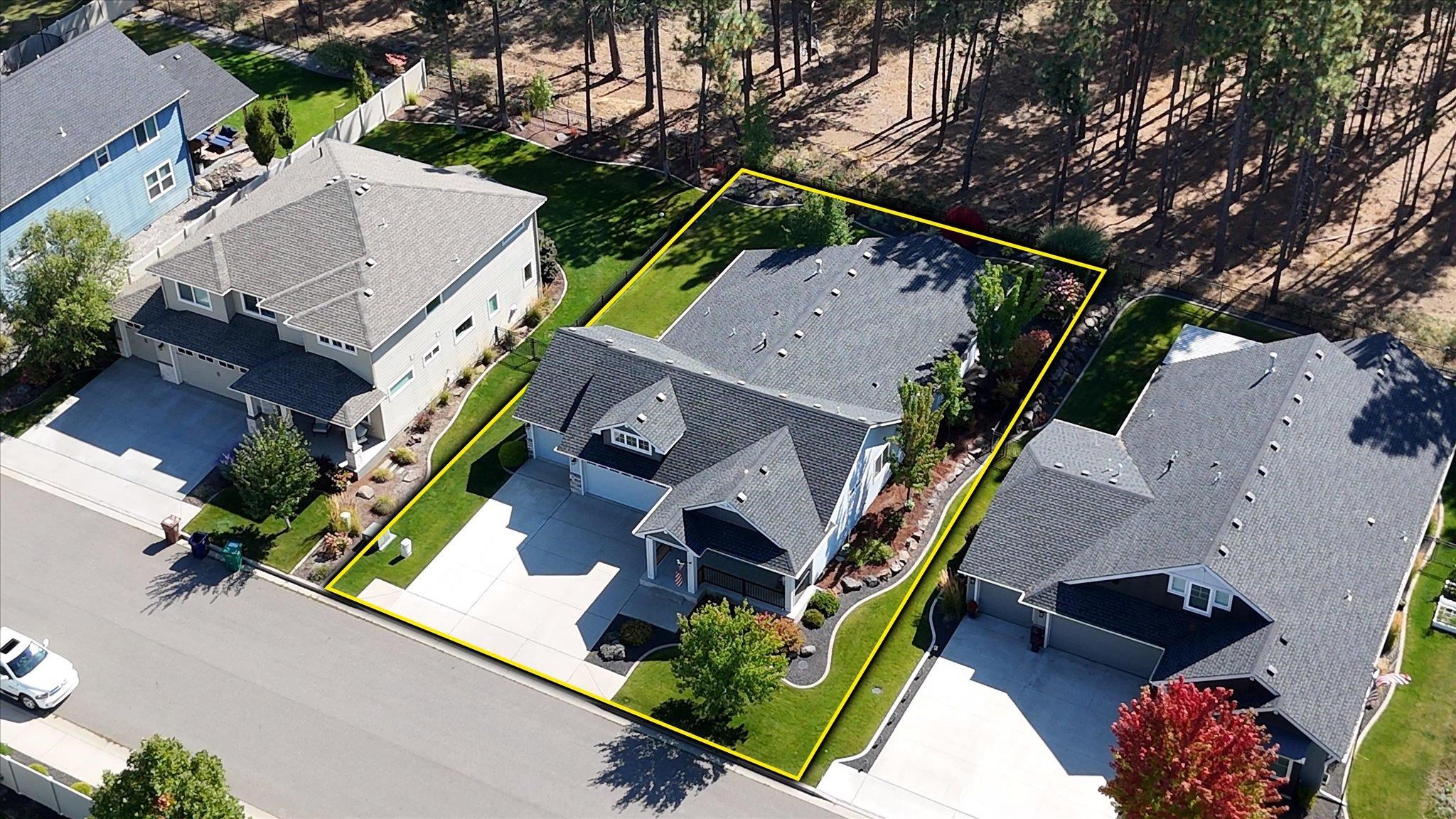 Aerial view of a home and landscaped lot outlined in yellow beside wooded land.