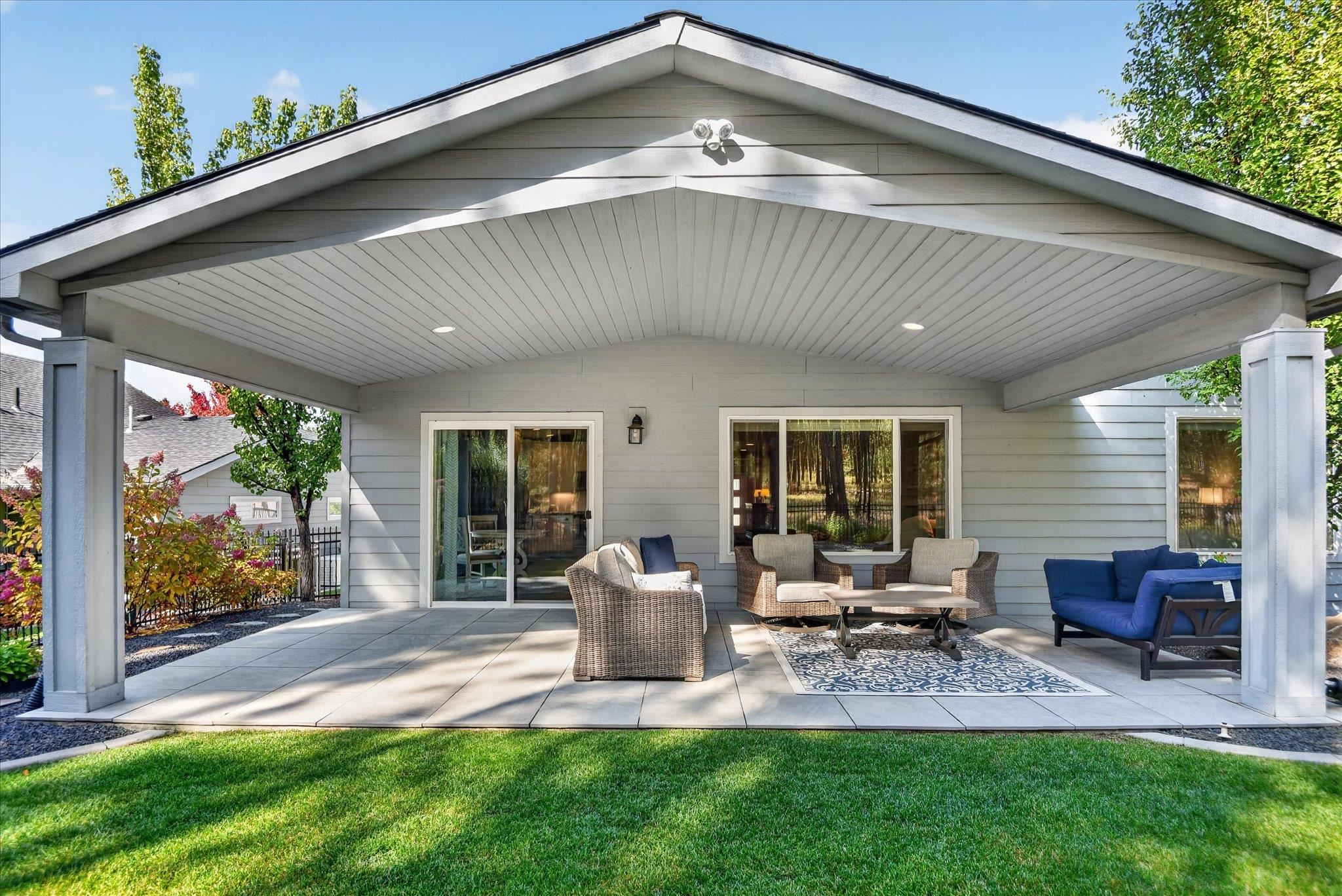 Covered backyard patio with wicker seating, blue loveseat, and patterned rug overlooking a green lawn.