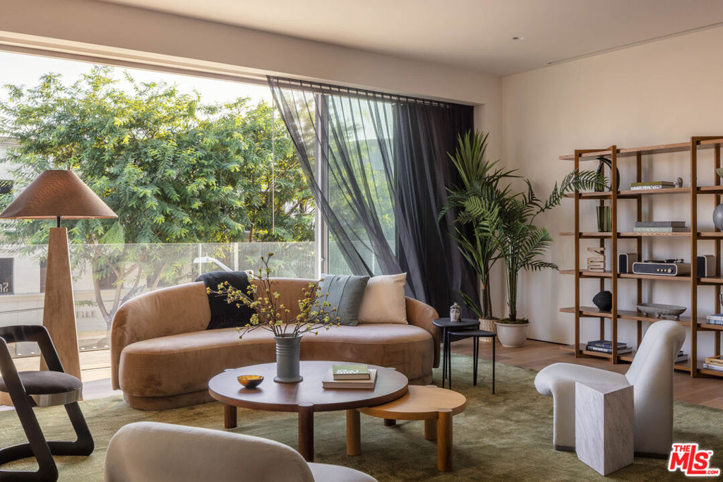 Staged living room with a curved tan sofa, round coffee table, and large windows overlooking leafy trees.