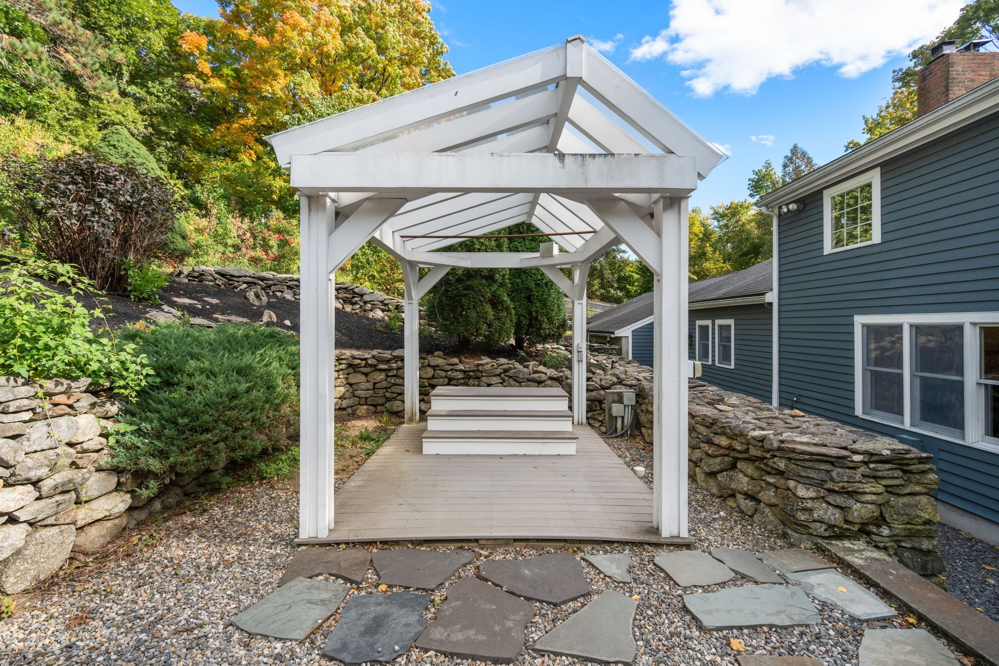 White timber pergola shelters a raised deck and steps, bordered by stone walls beside a blue-gray house.