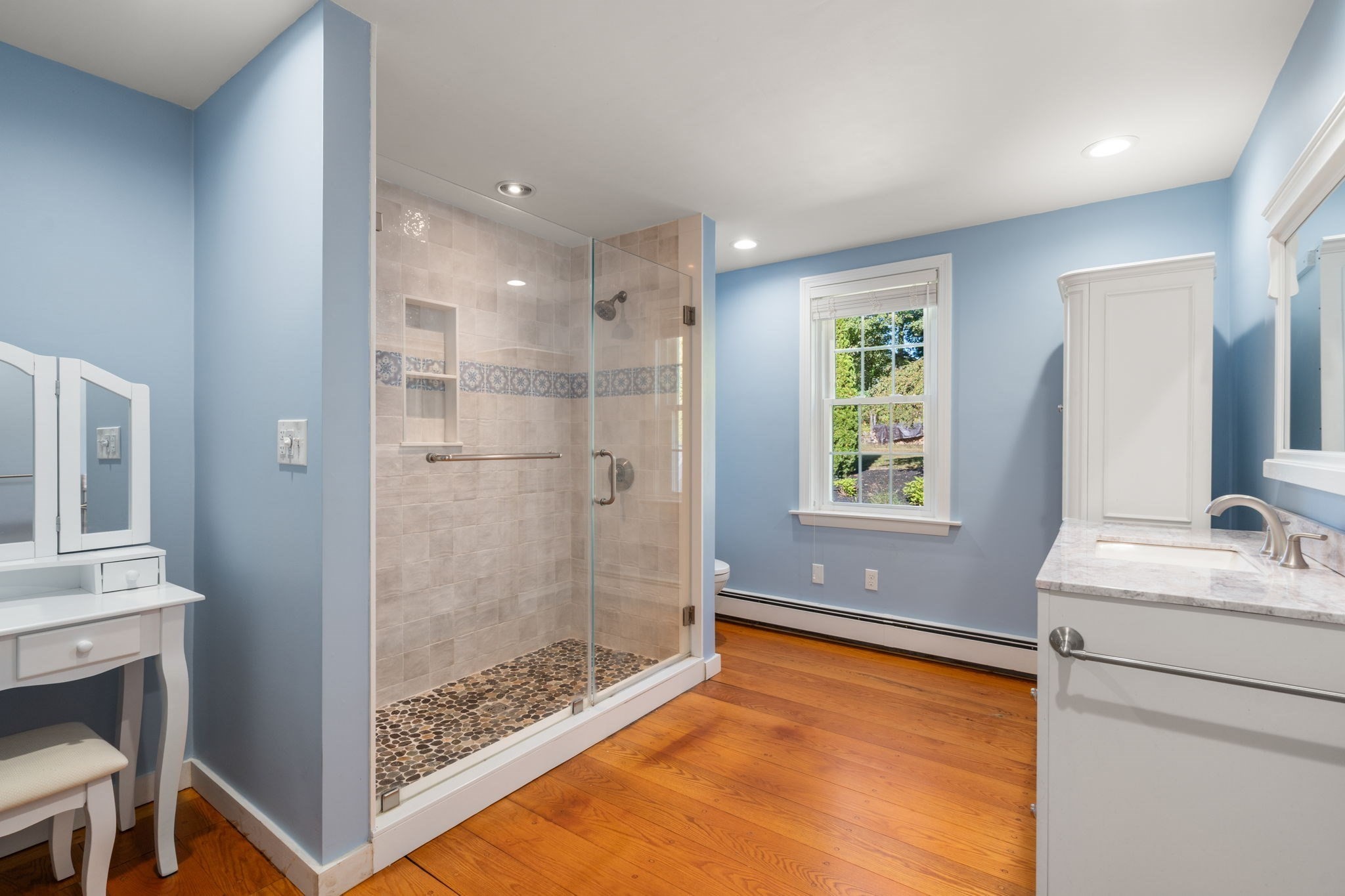 Bathroom with a spacious glass shower, pebble floor, blue walls, and warm hardwood flooring.