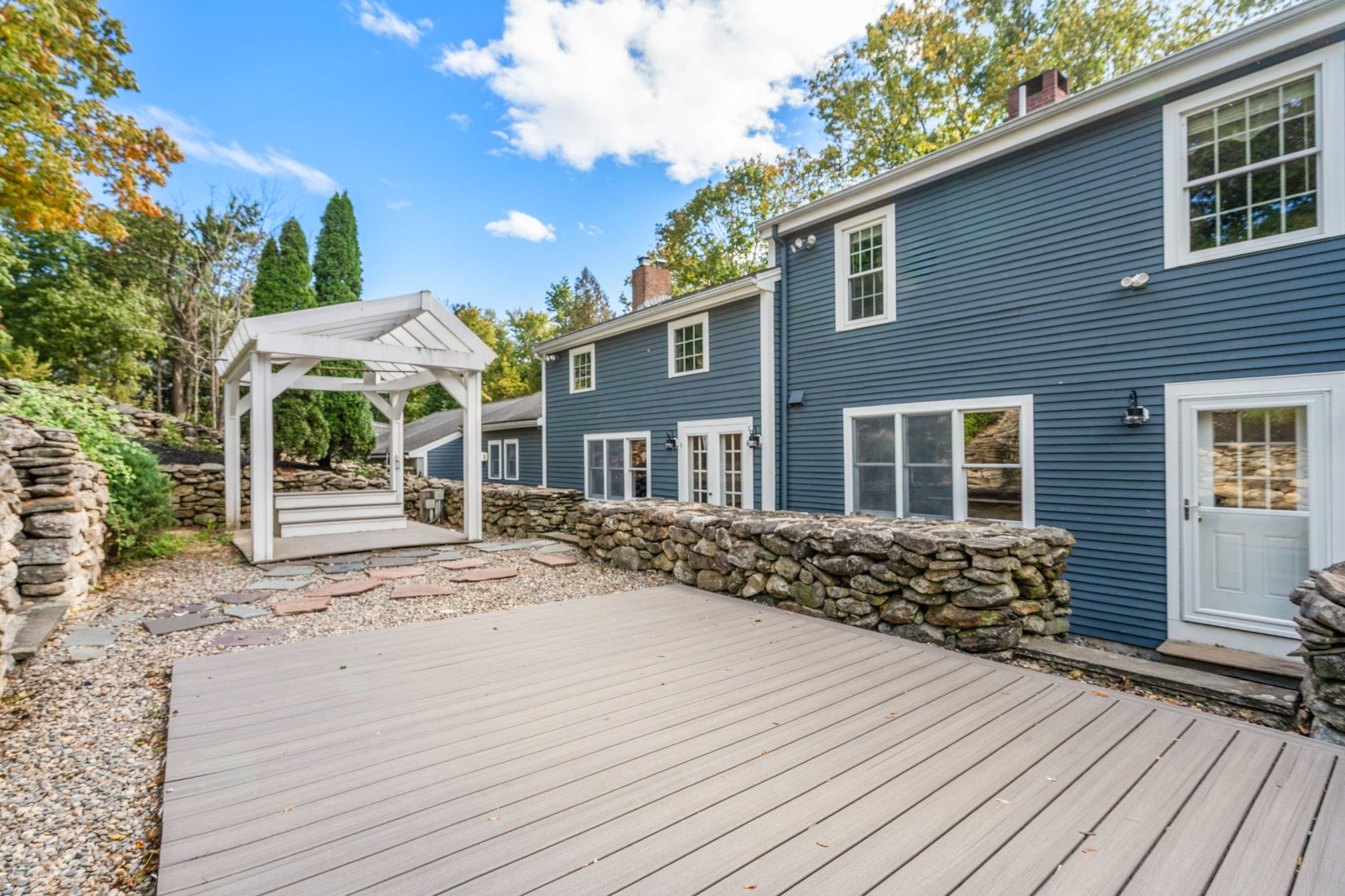 Blue house beside a broad deck, white pergola, and low stone walls.