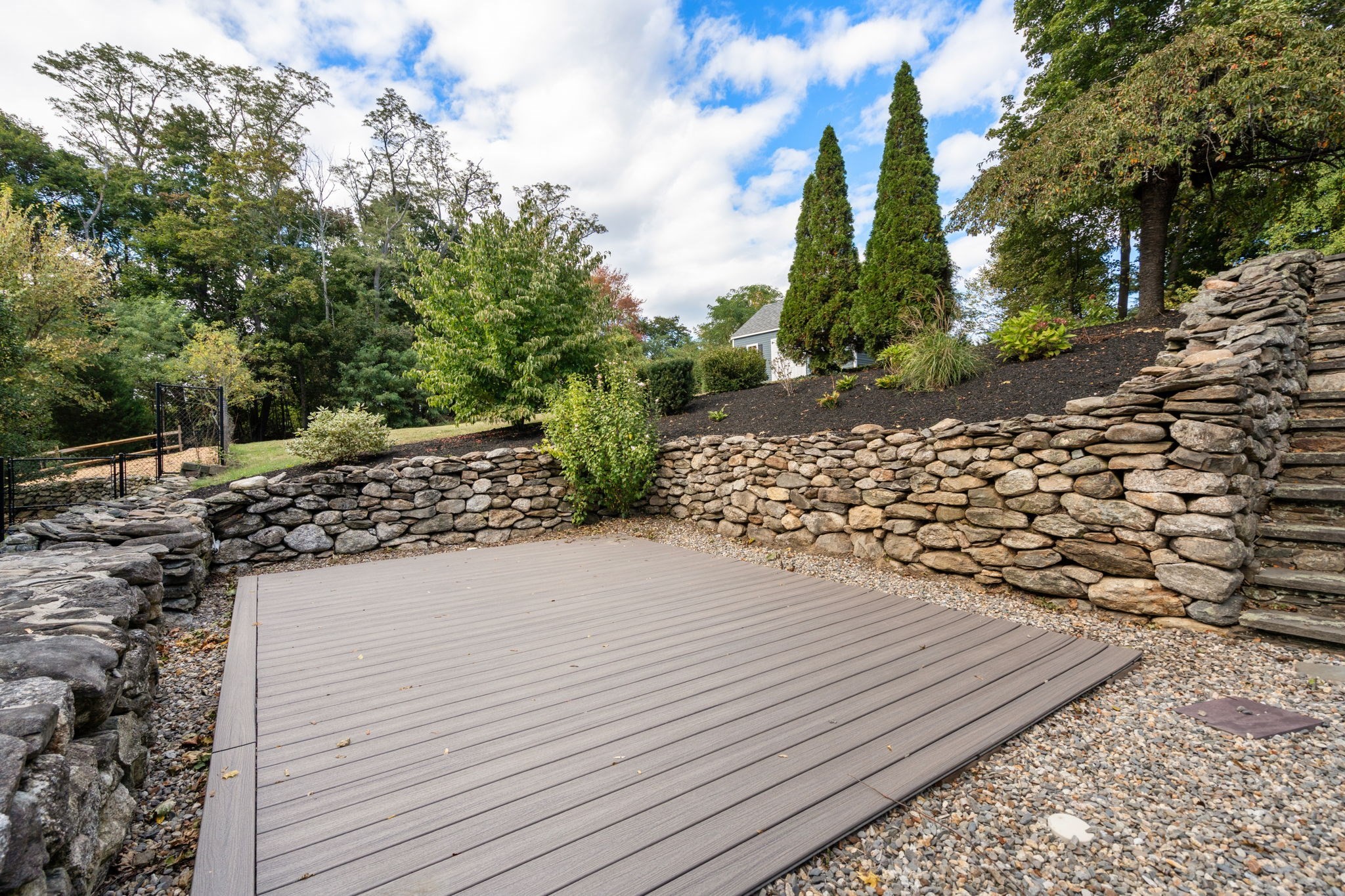 Outdoor patio with a broad composite deck, stone retaining walls, and landscaped evergreen slope.