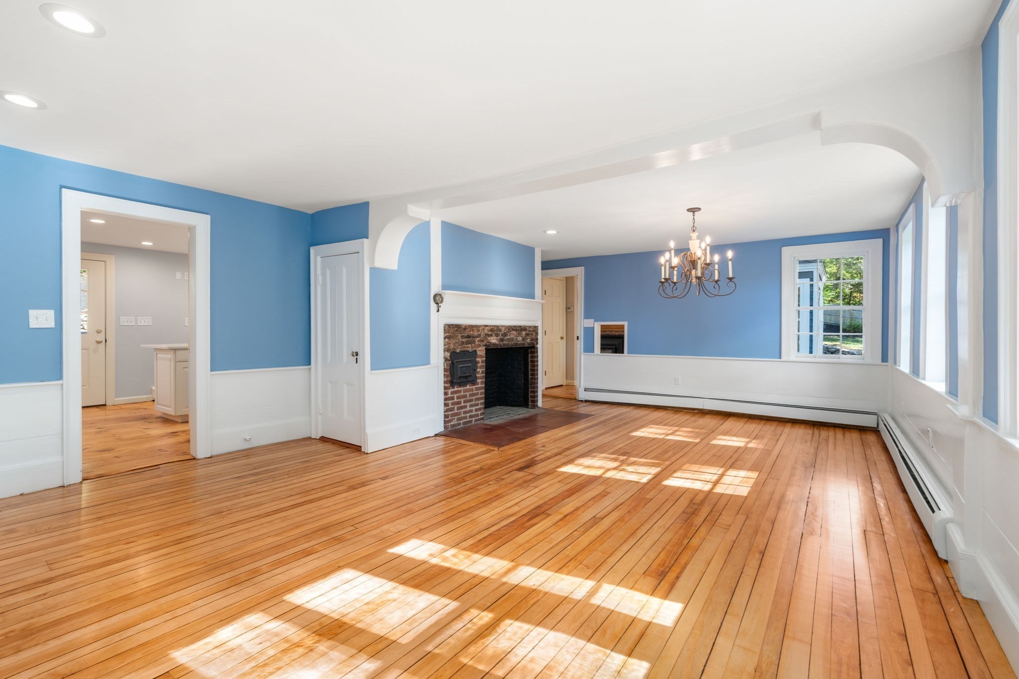 Vacant living and dining room with blue walls, hardwood floors, a brick fireplace, and large windows.