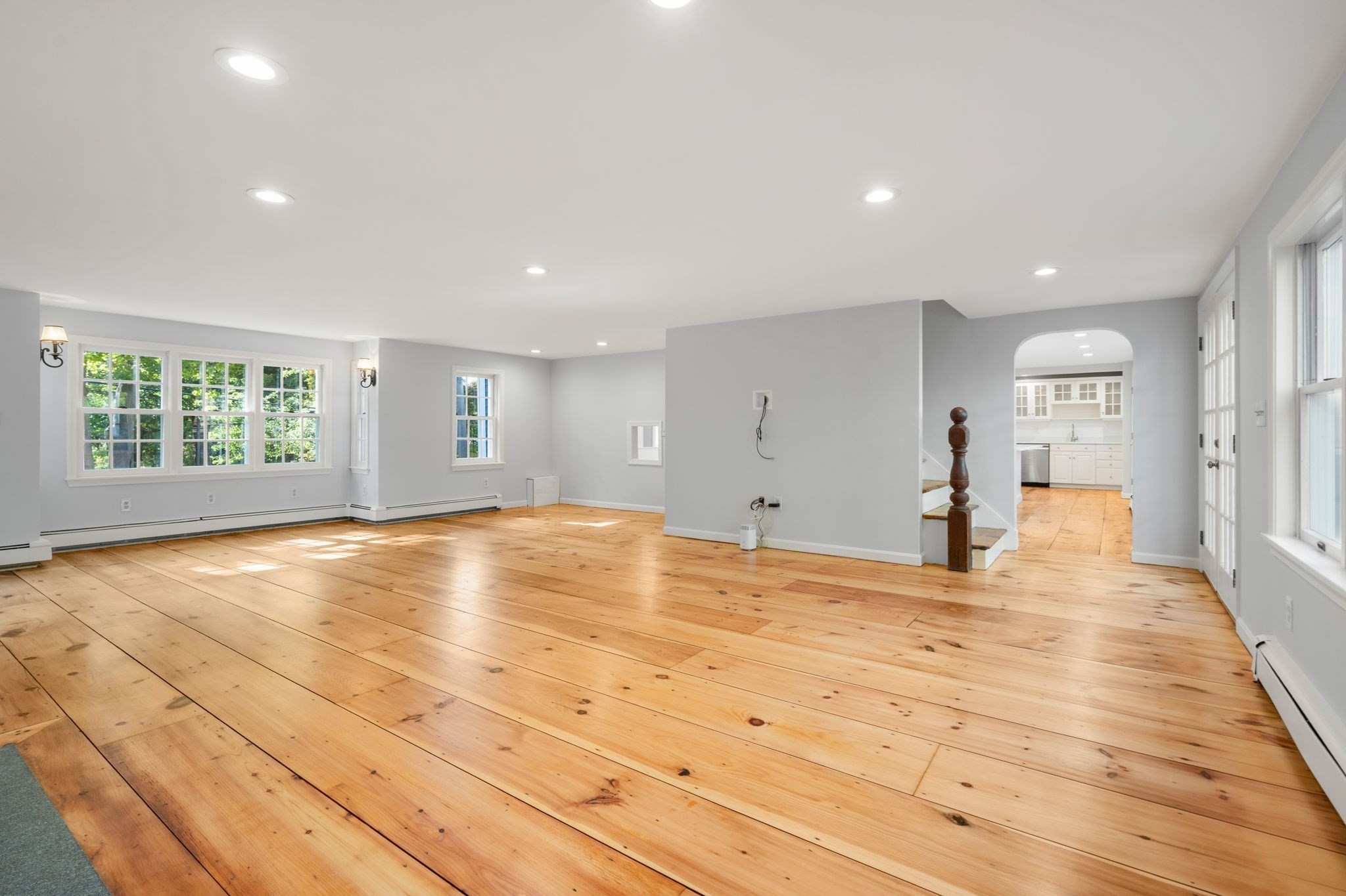 Vacant living room with wide-plank wood floors, large windows, and an arched opening to the kitchen.