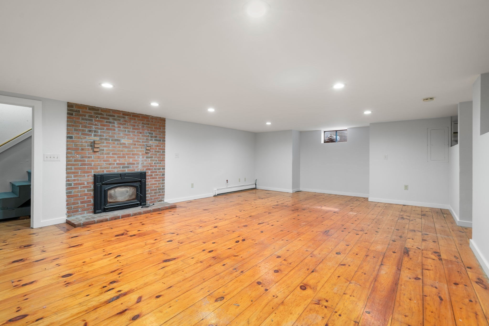 Empty basement living area with wide-plank wood floors and a black fireplace against a brick wall.
