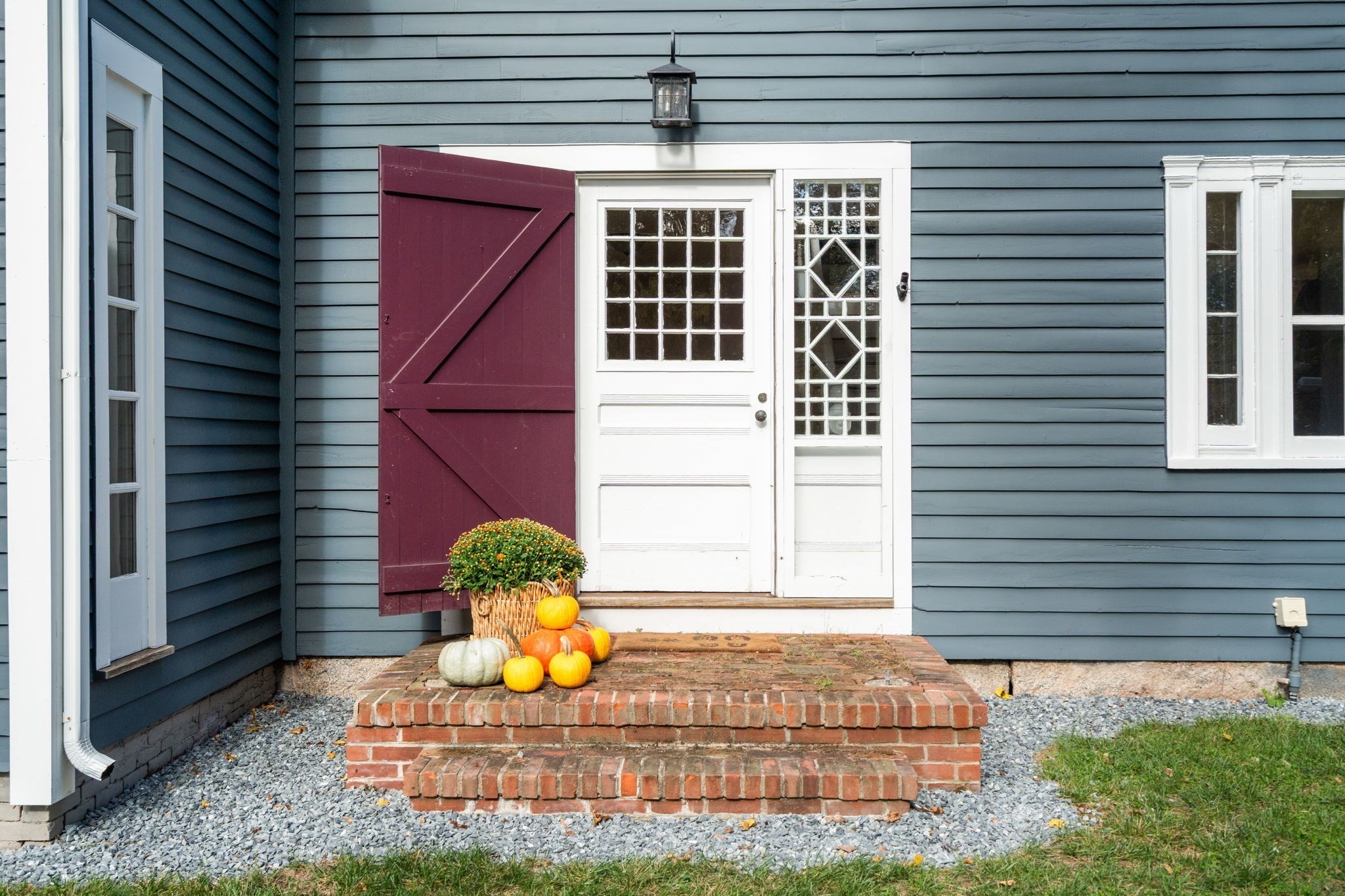 House entrance with a white divided-light door, burgundy shutter, brick steps, and autumn pumpkins.
