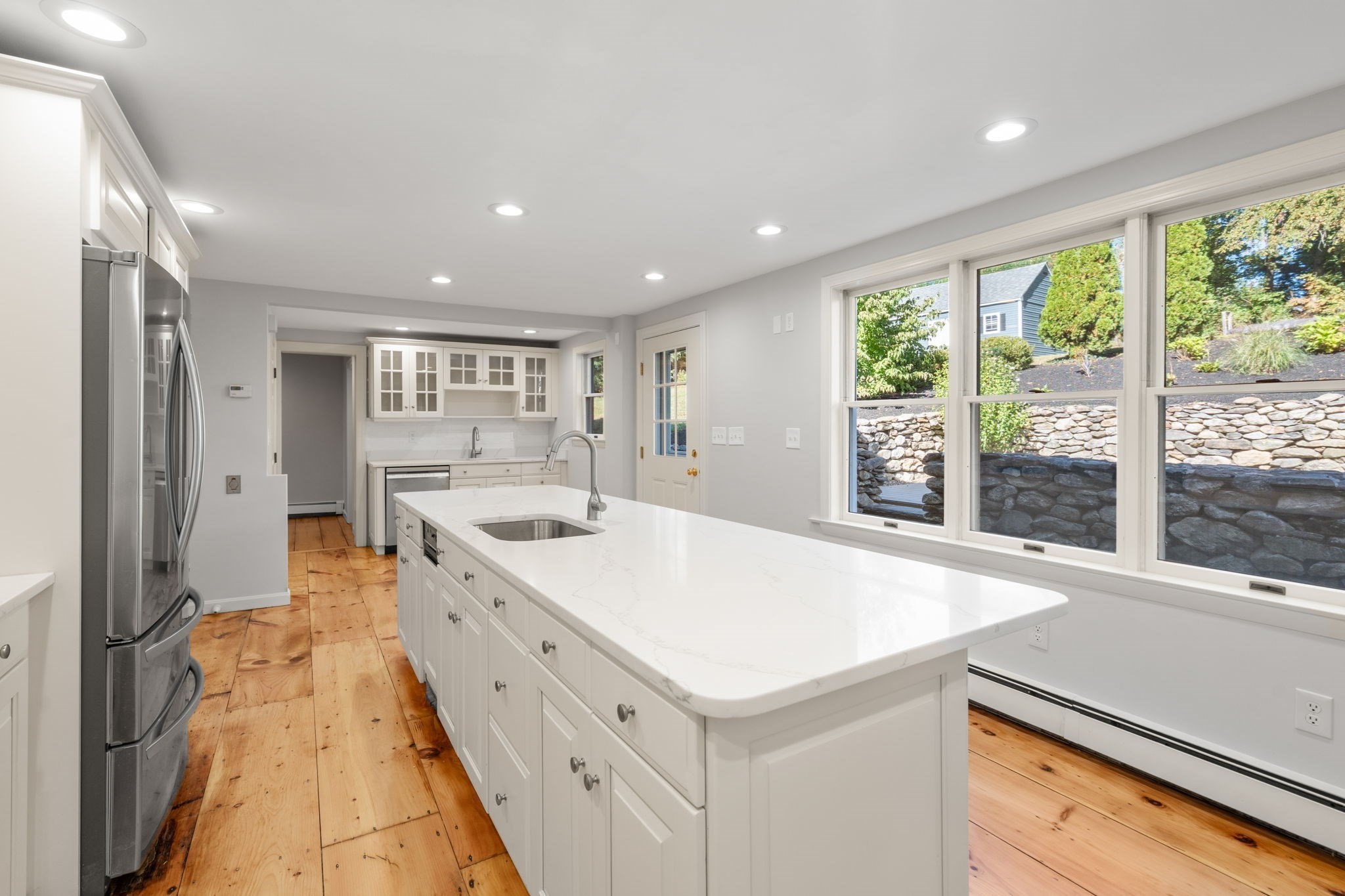 Bright white kitchen with a large quartz island, stainless steel refrigerator, and windows above a stone retaining wall.