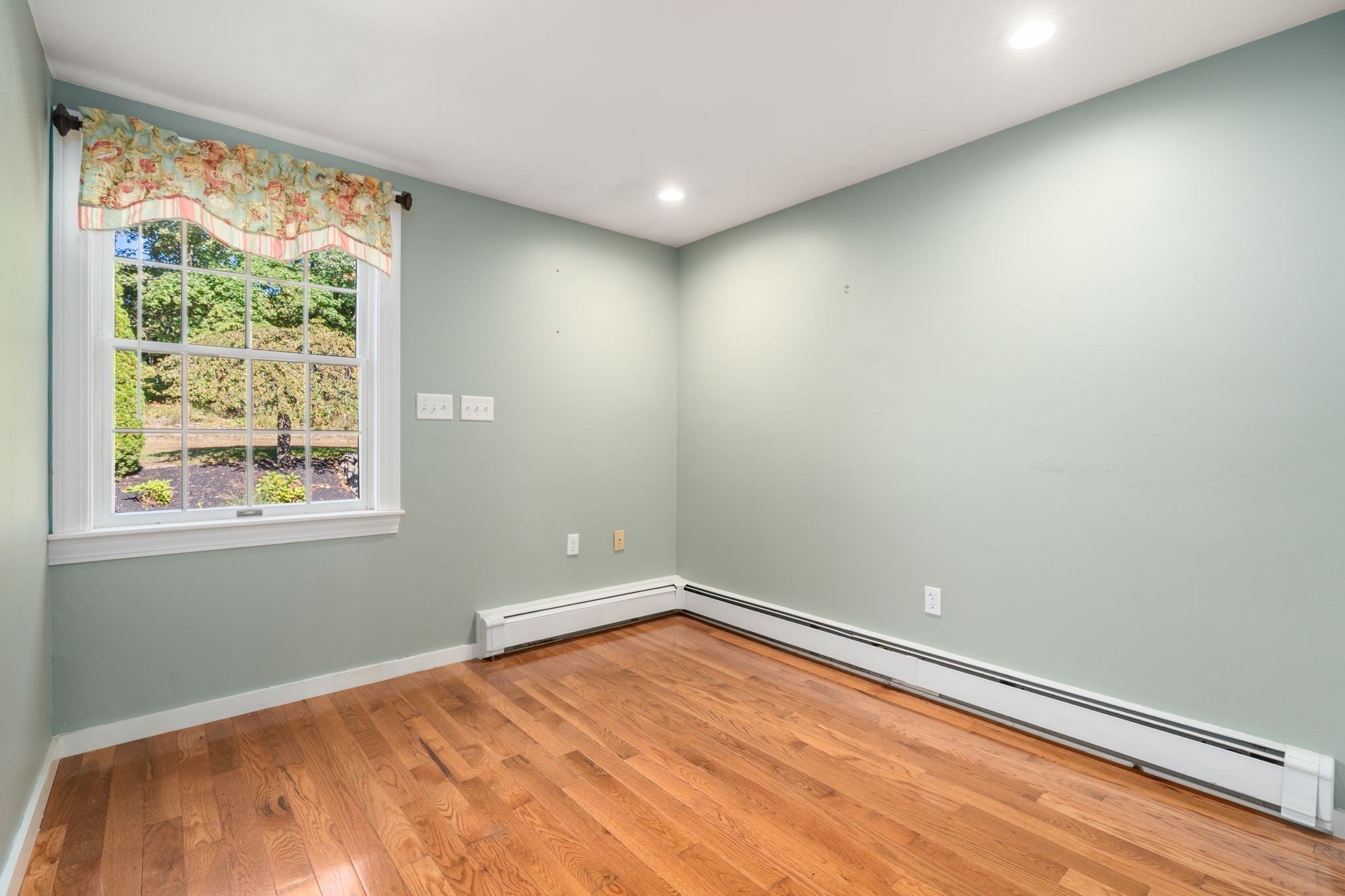 Empty room with hardwood floors, pale green walls, and a large window with a floral valance.