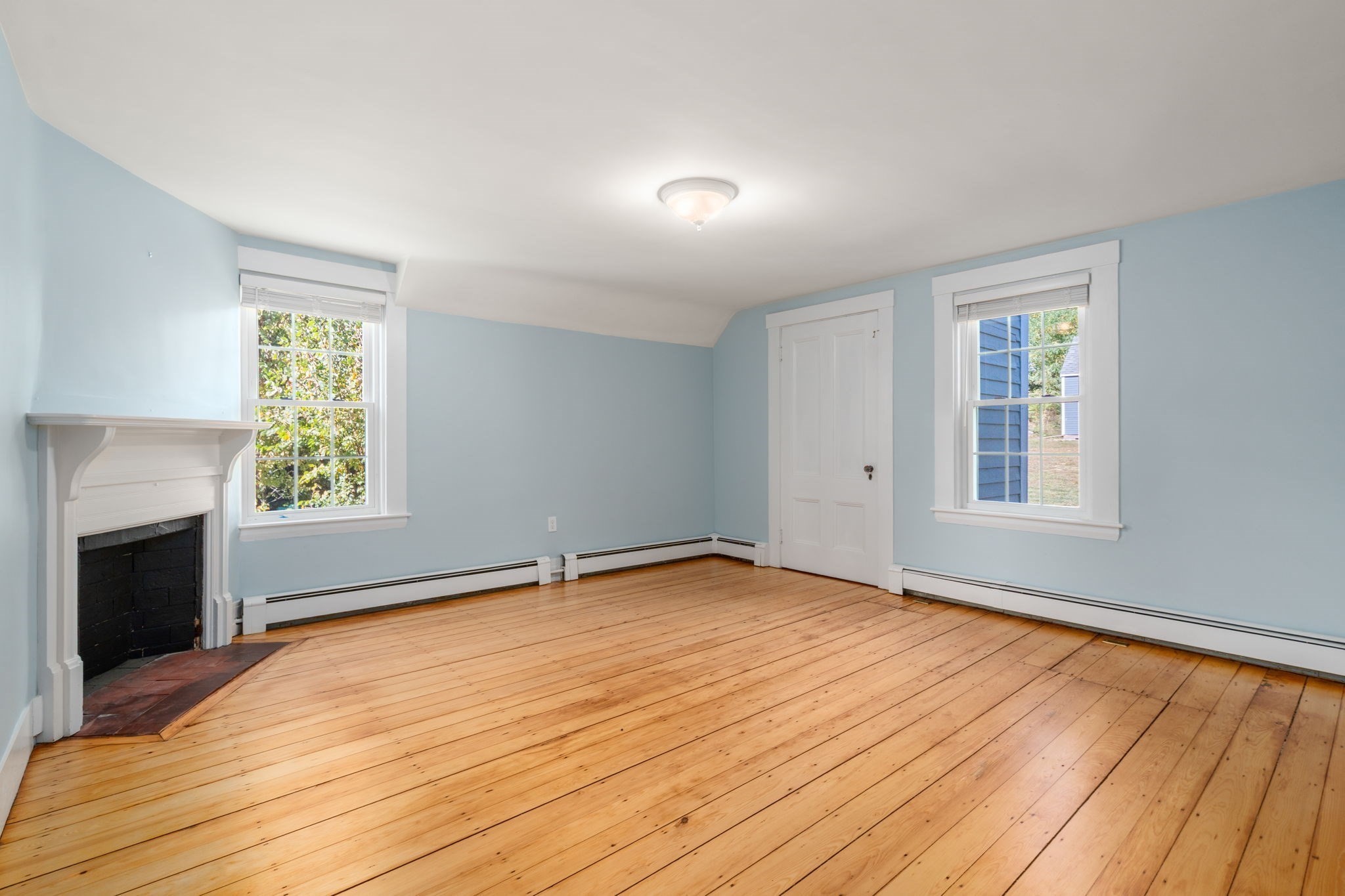 Empty pale blue room with hardwood floors, a white fireplace, and two windows.