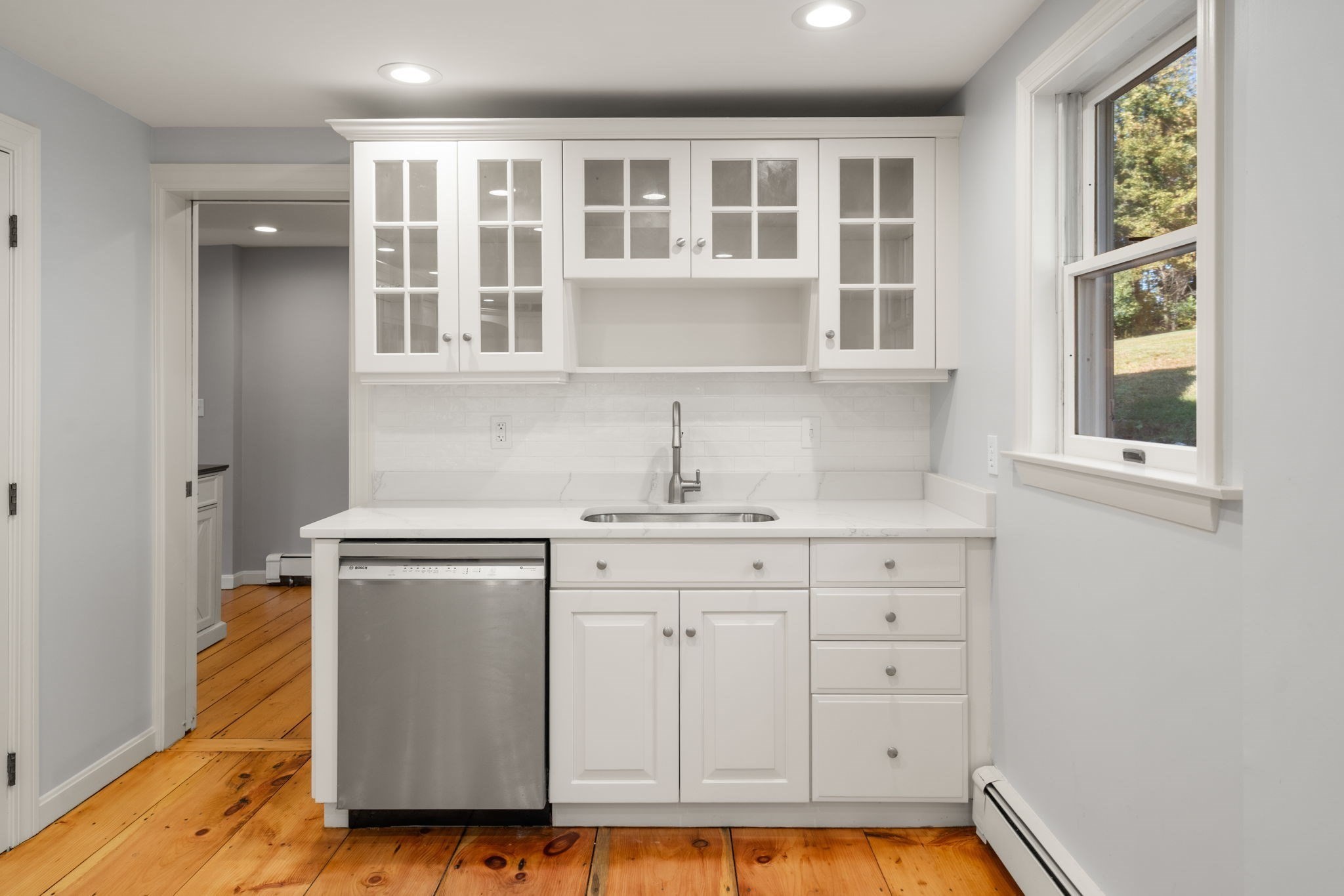 Kitchen with white cabinets, stainless steel dishwasher, and warm hardwood floors beside a large window.