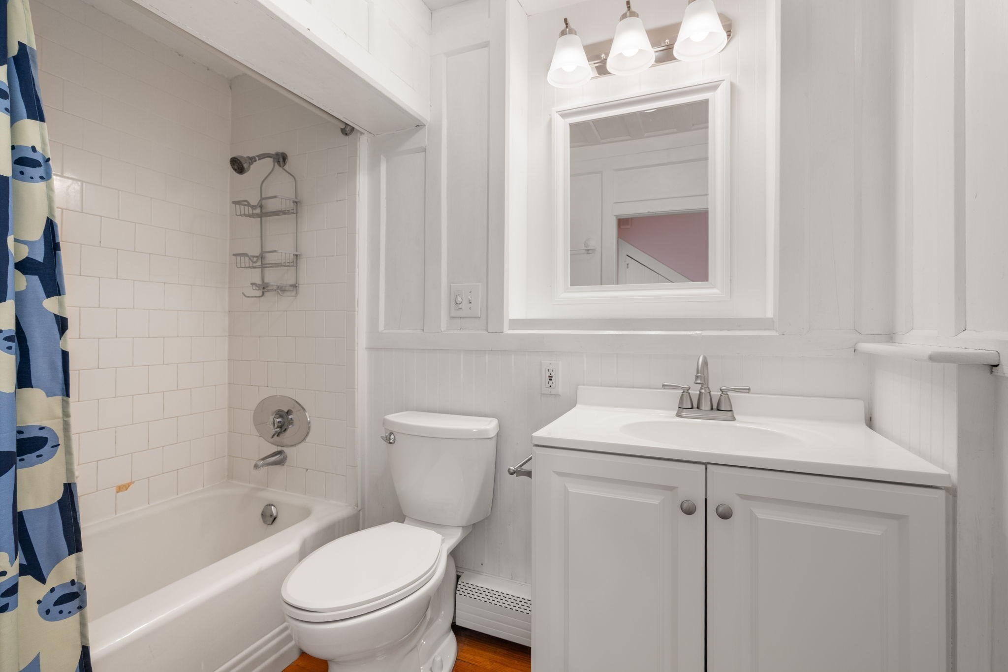 Bathroom with a white tiled tub and shower, blue patterned curtain, and white vanity.