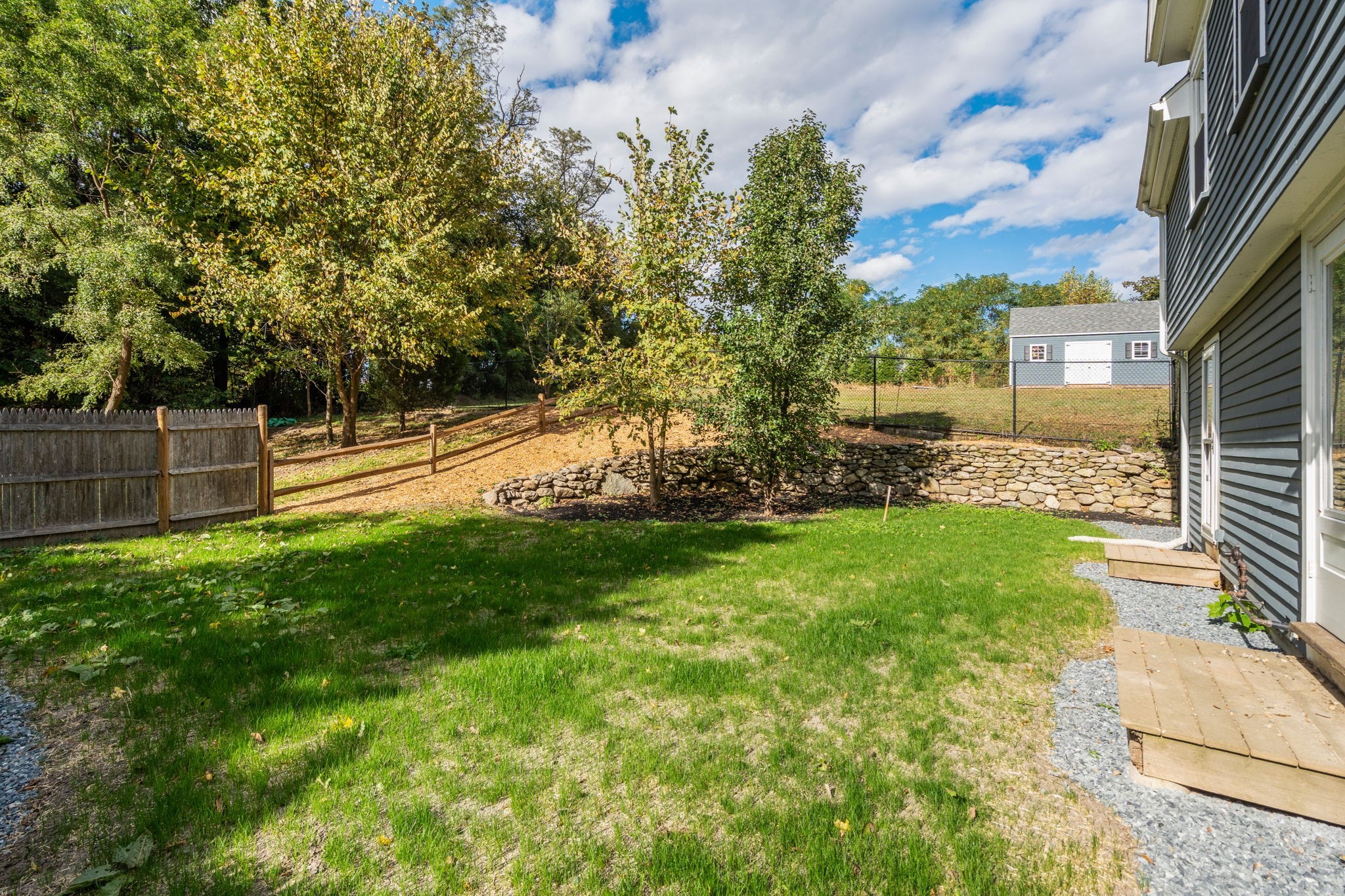 Backyard with a green lawn, mature trees, stone retaining wall, and fenced outbuilding.