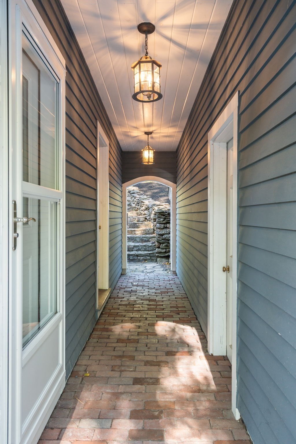 Covered passage with a brick floor, blue siding, hanging lanterns, and stone steps.