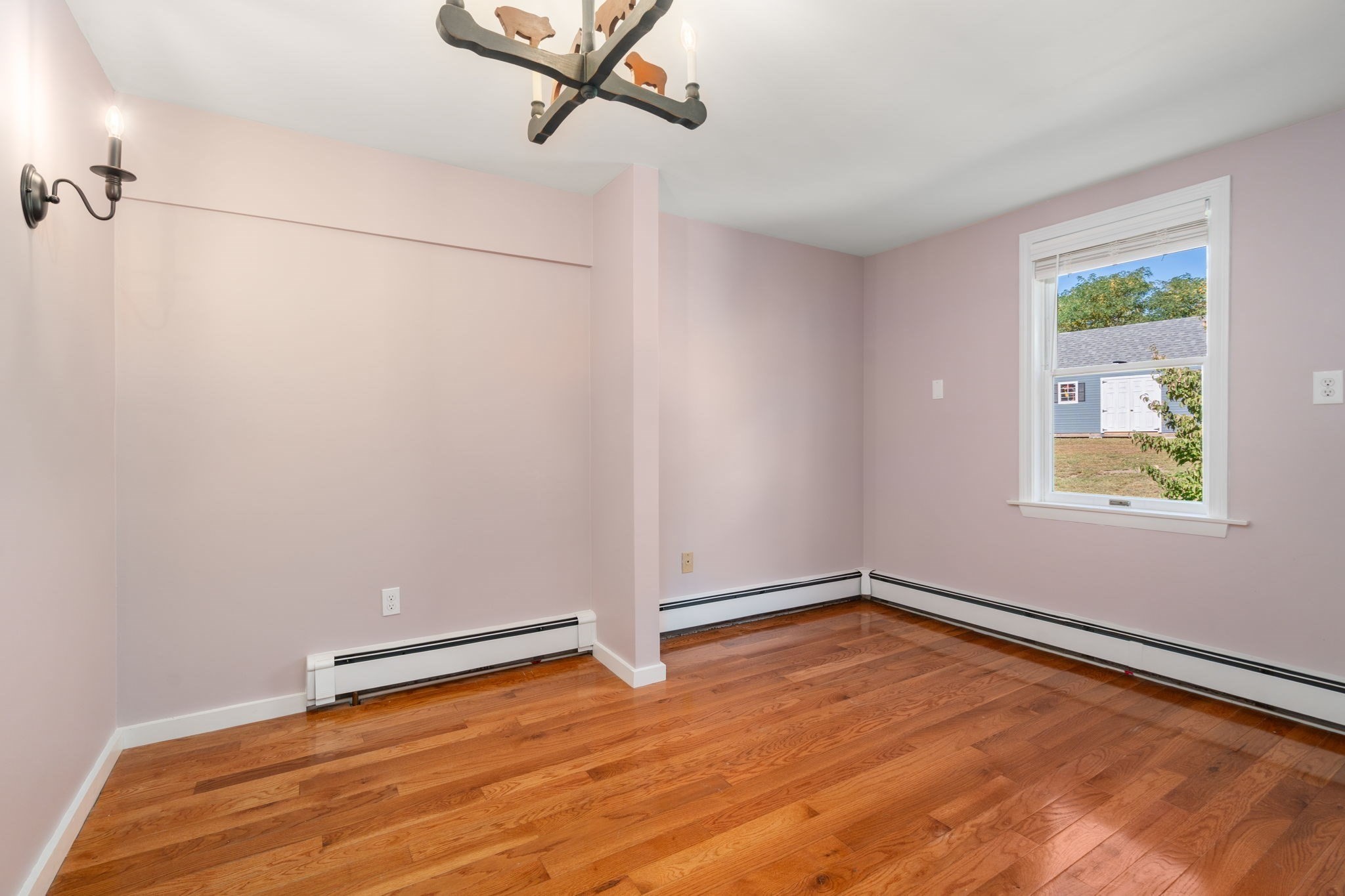Empty room with pale pink walls, polished hardwood floors, and a window overlooking a small outbuilding.