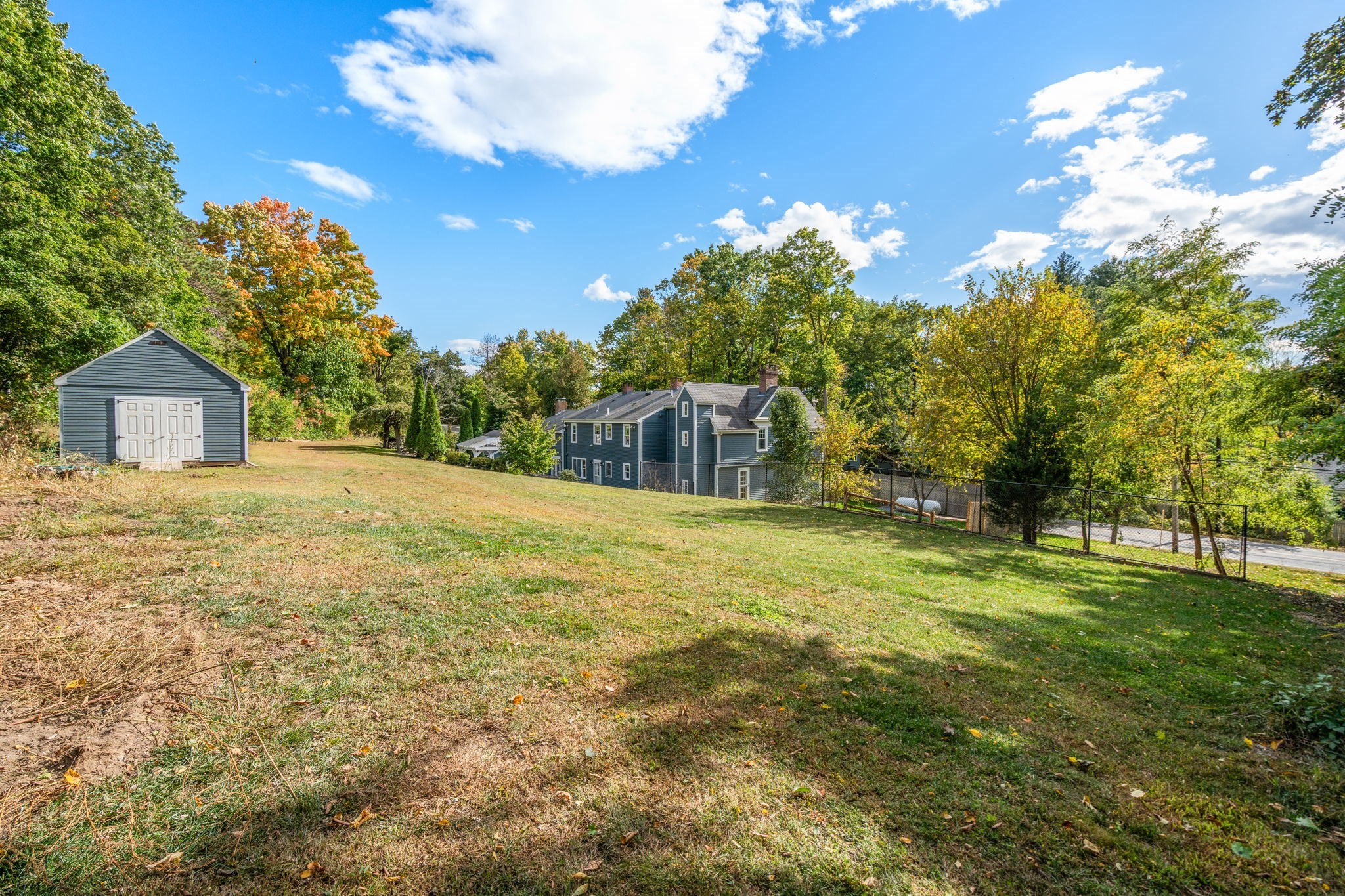 Sloping lawn with a blue-gray house and detached garage among mature autumn trees.