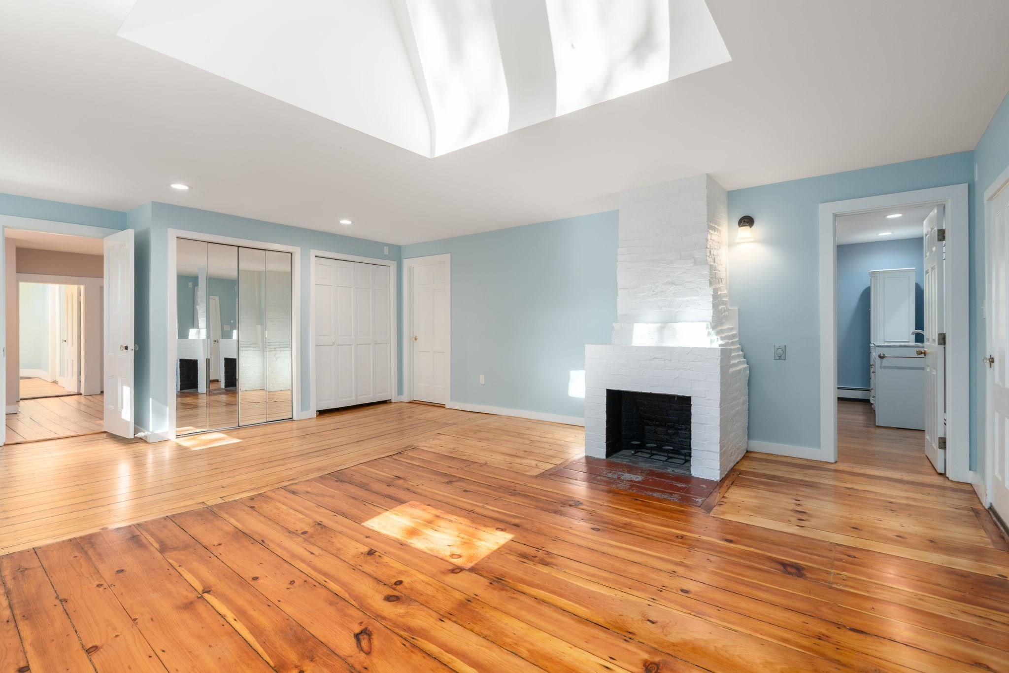 Empty room with wide plank wood floors, a white brick fireplace, and a large skylight.