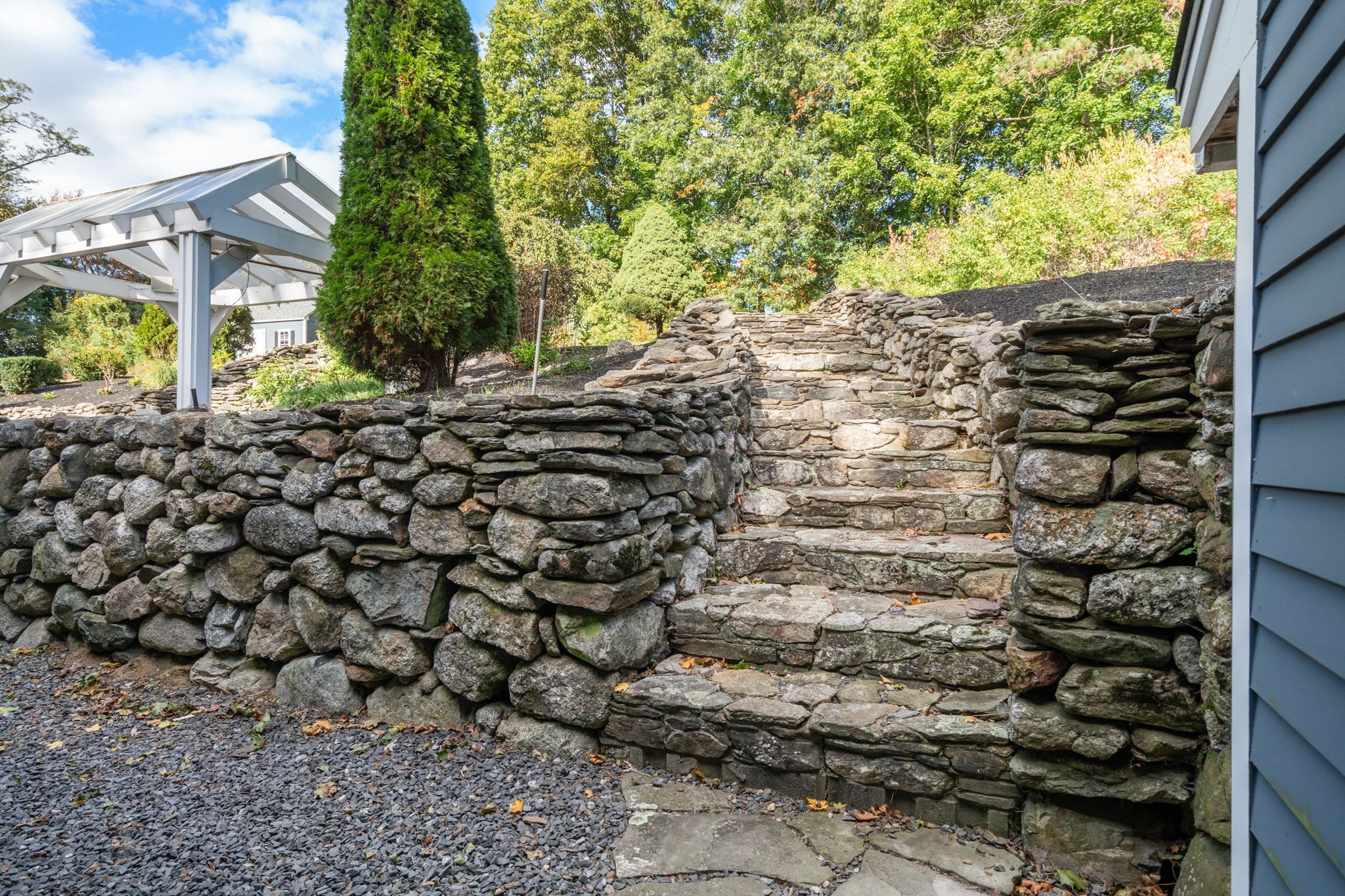 Stone steps rise between stacked-stone retaining walls beside a white pergola and blue house.