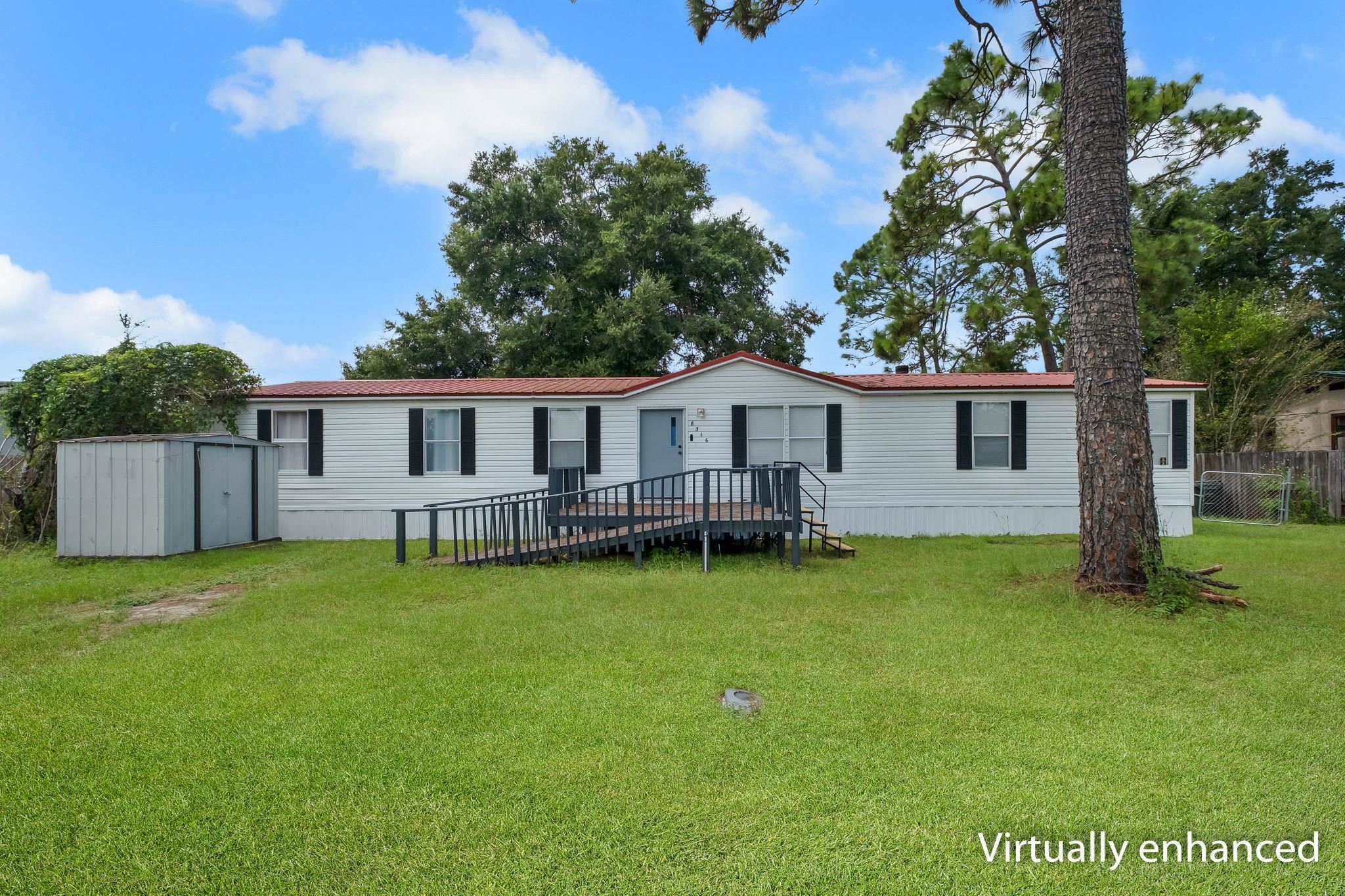 White single-story home with a red metal roof, raised ramp, and gray storage shed on a tree-lined lawn.