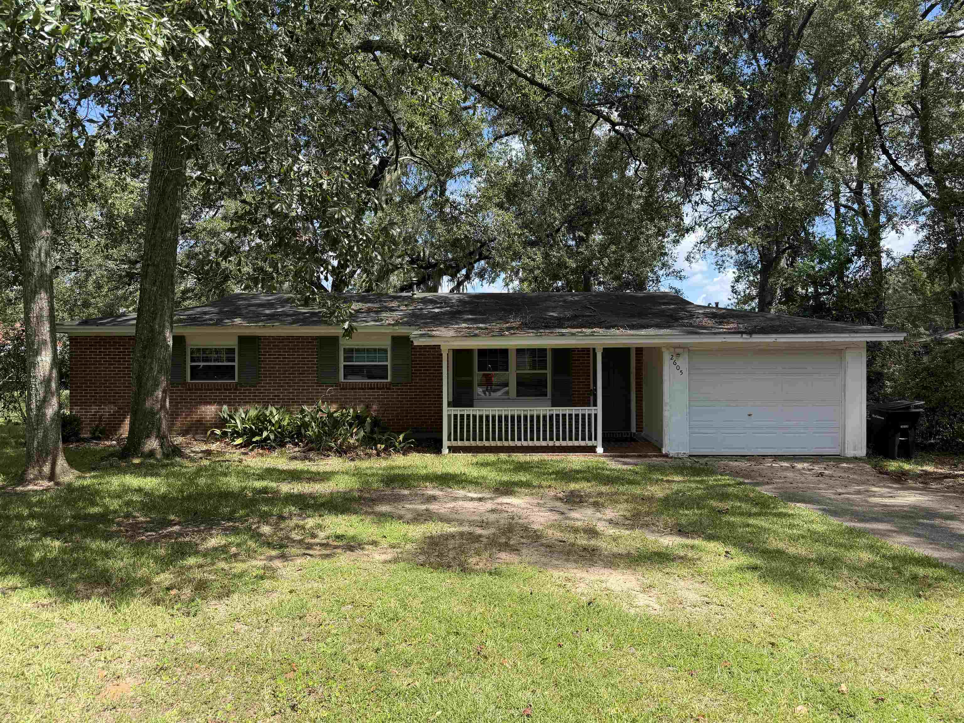 Single-story brick house with a covered porch and attached white garage beneath mature trees.