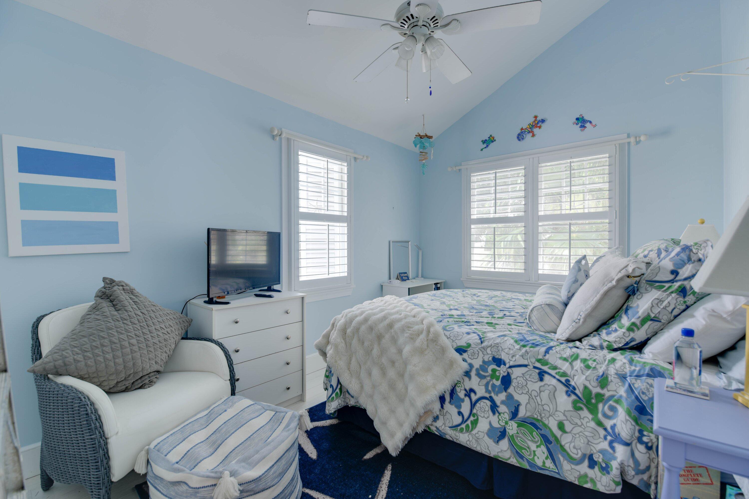 Bedroom with pale blue walls, a patterned bedspread, white shutters, and a wicker armchair.