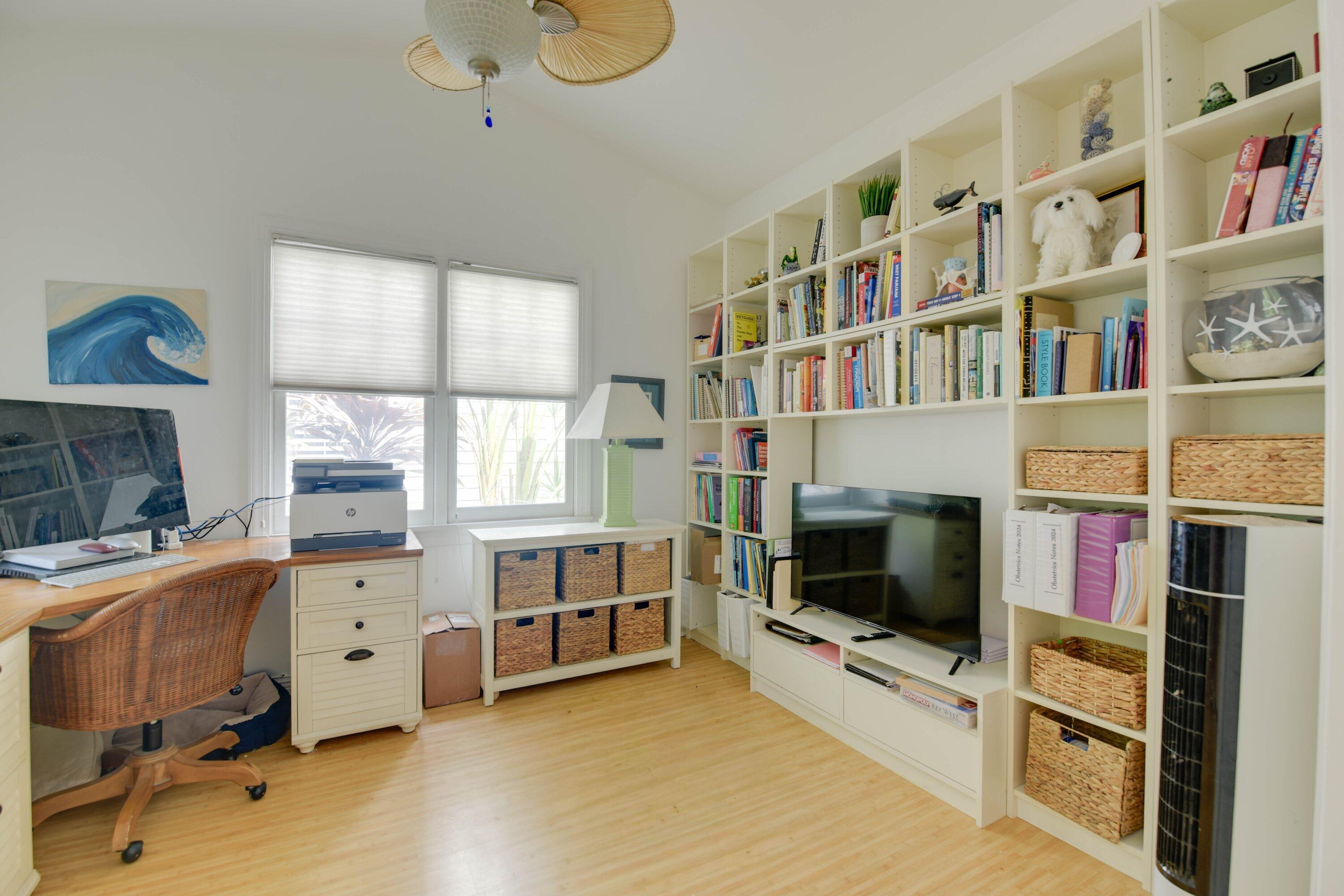 Home office with built-in white bookshelves, woven baskets, a television, and a desk by two windows.