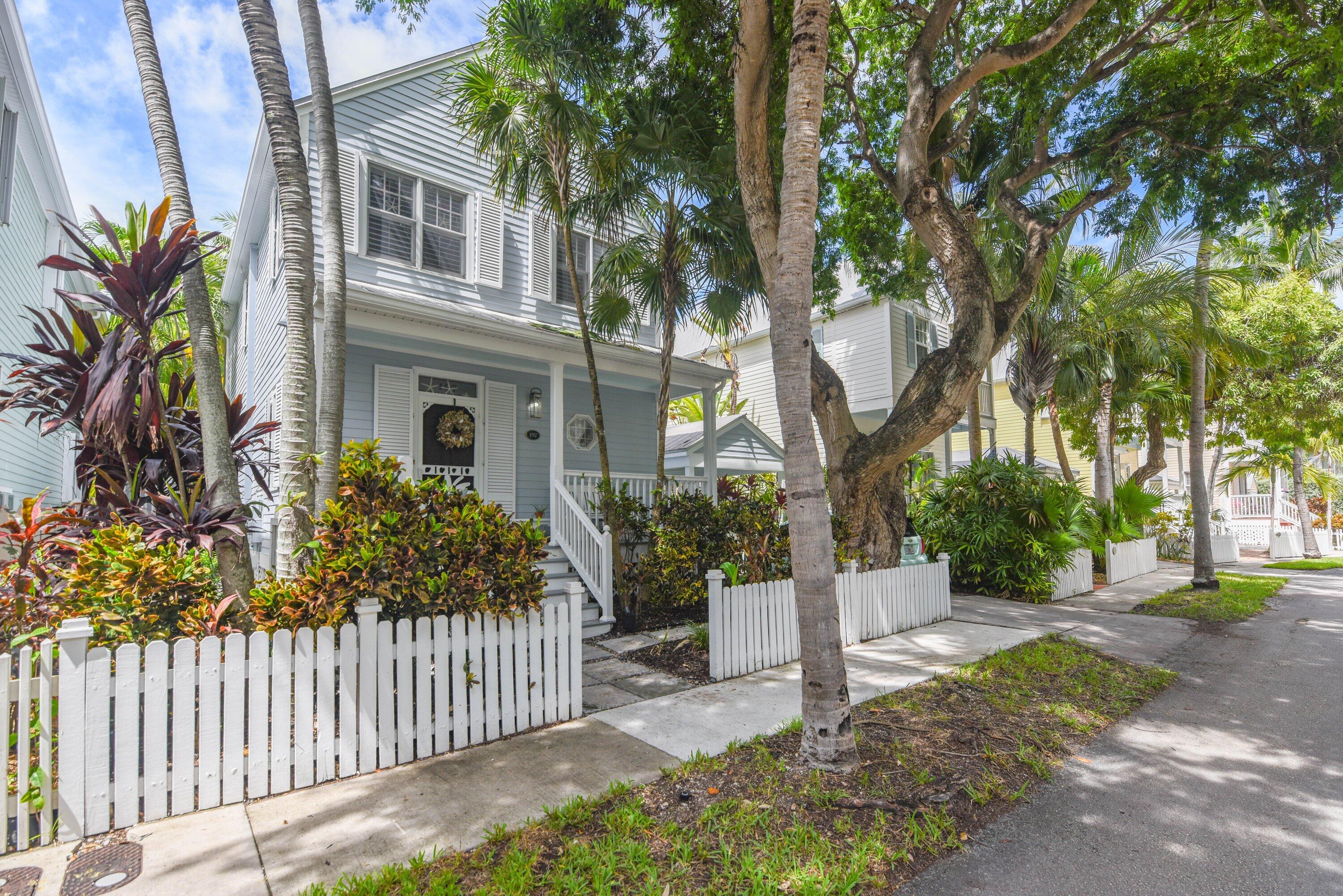Pale blue two-story house with a white picket fence, palm trees, and a shaded front porch.