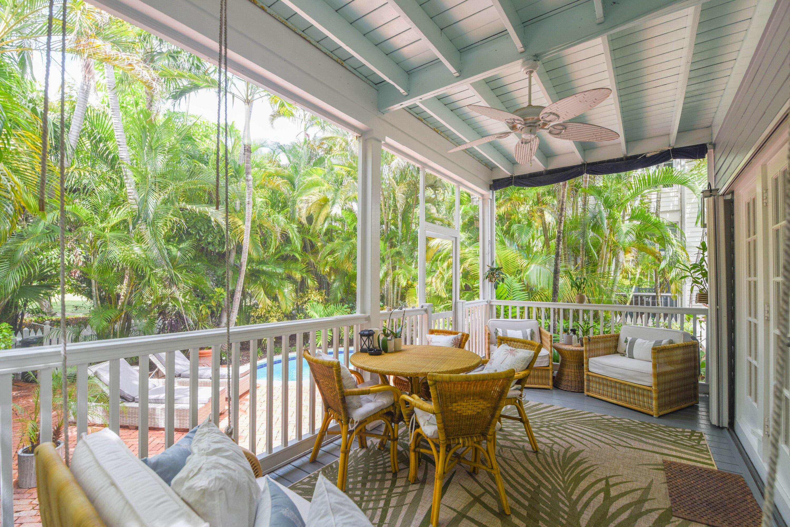 Covered porch with wicker dining and lounge seating, overlooking tropical palms and a glimpse of the pool.