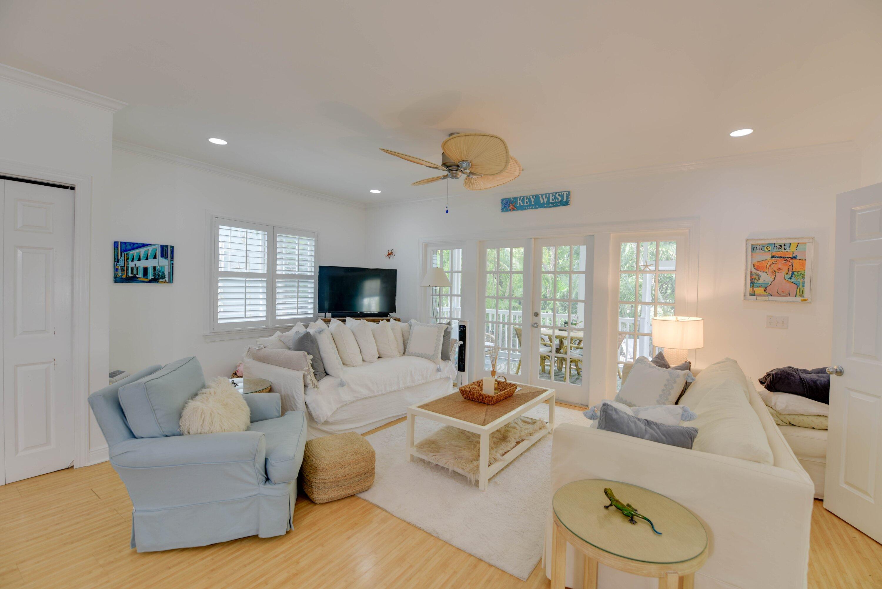 Furnished living room with white sofas, a pale blue armchair, and French doors opening to a leafy porch.