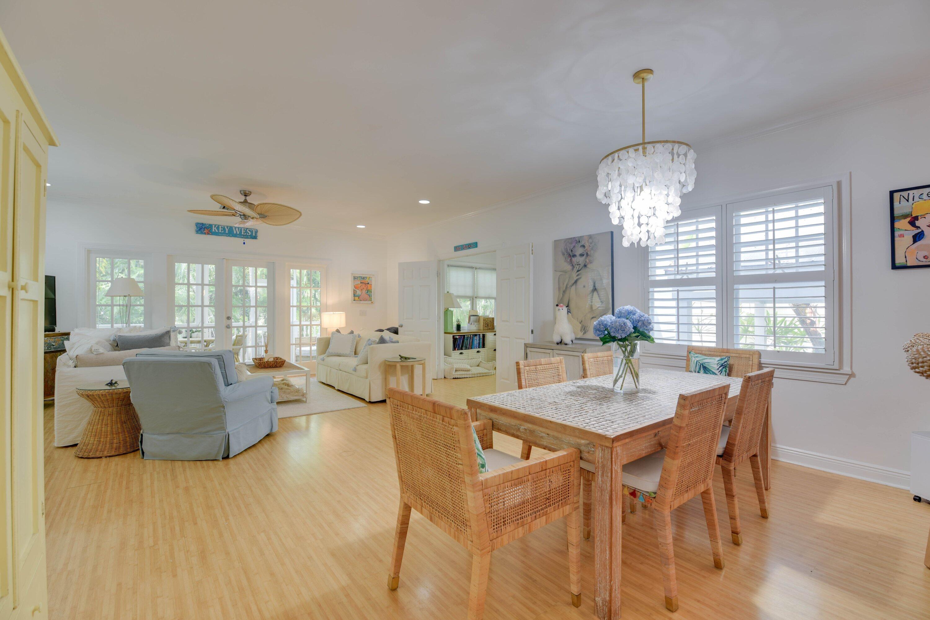 Open living and dining room with white sofas, woven furniture, shuttered windows, and a shell chandelier.