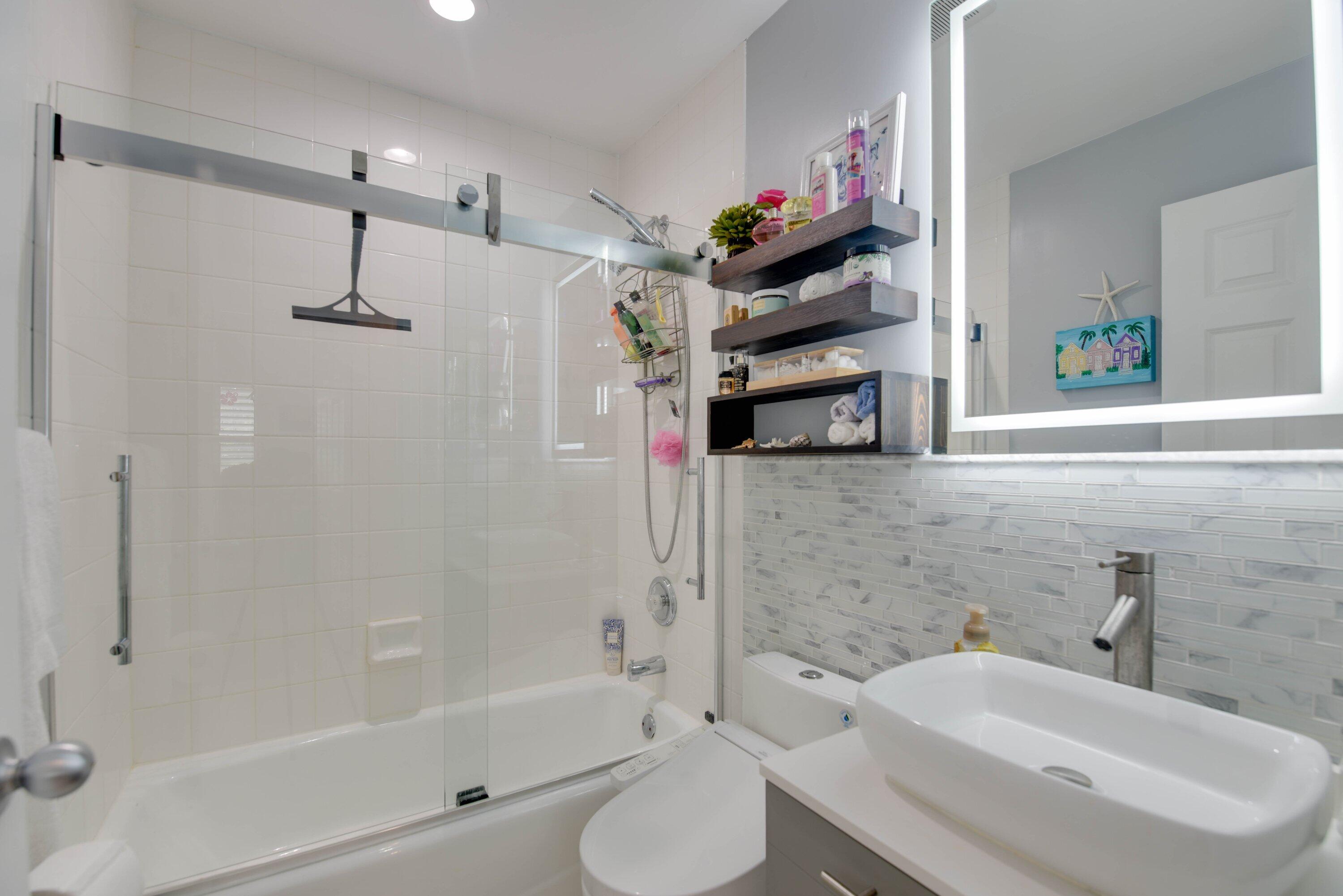 Modern bathroom with glass-enclosed bathtub, vessel sink, illuminated mirror, and floating shelves.