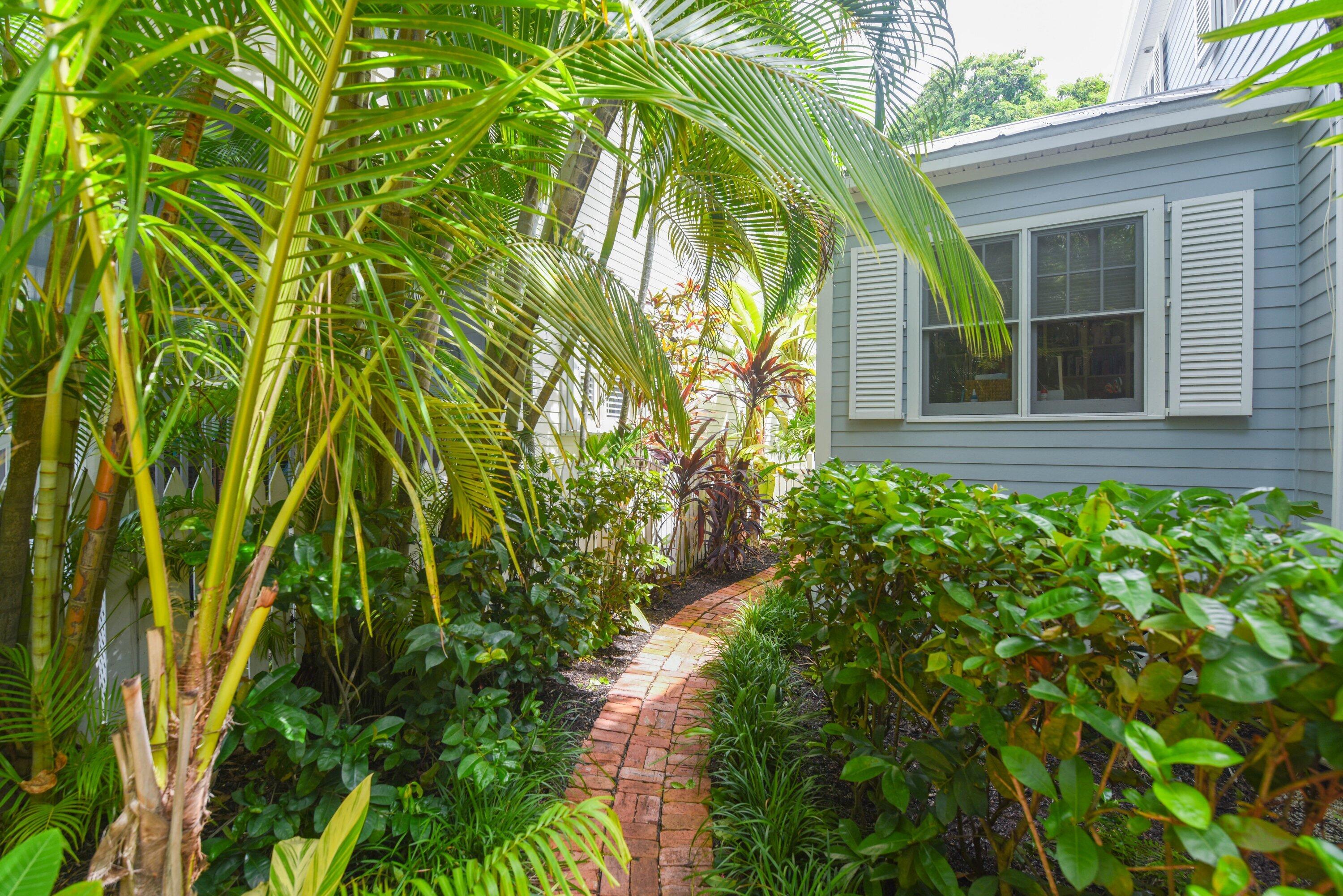 Blue-gray house beside a curved brick path framed by lush palms and tropical shrubs.