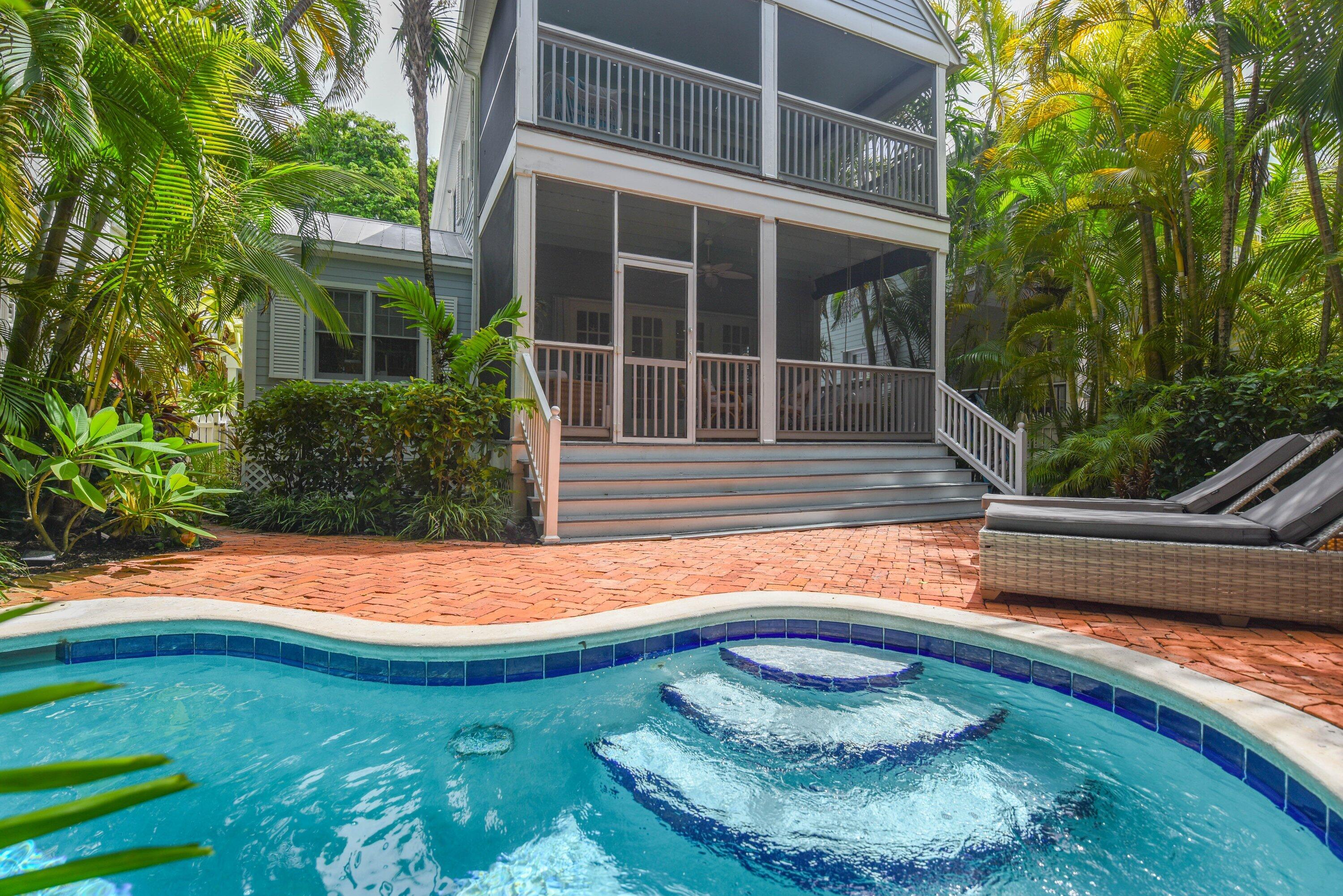 Backyard pool beside a brick patio, screened porch, and palm-filled landscaping.