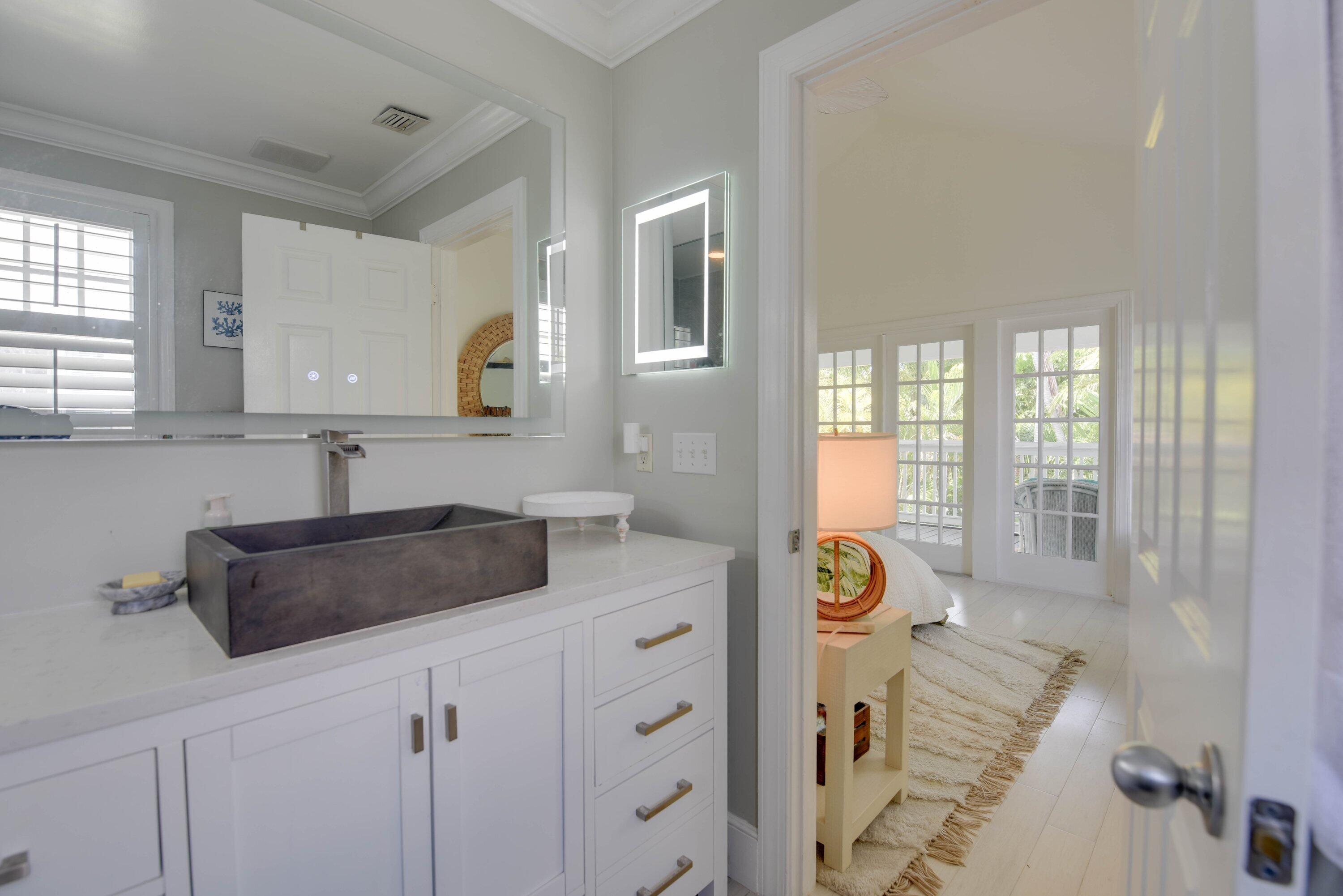 Bathroom vanity with dark vessel sink and lit mirror, looking into a bedroom with balcony doors.