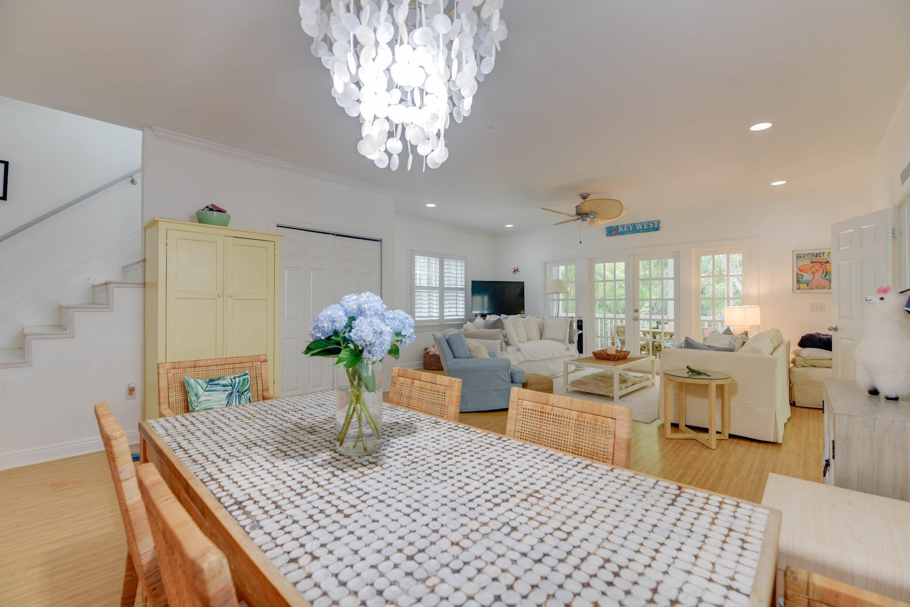 Open-plan dining and living room with a patterned table, blue hydrangeas, and glass doors to greenery.
