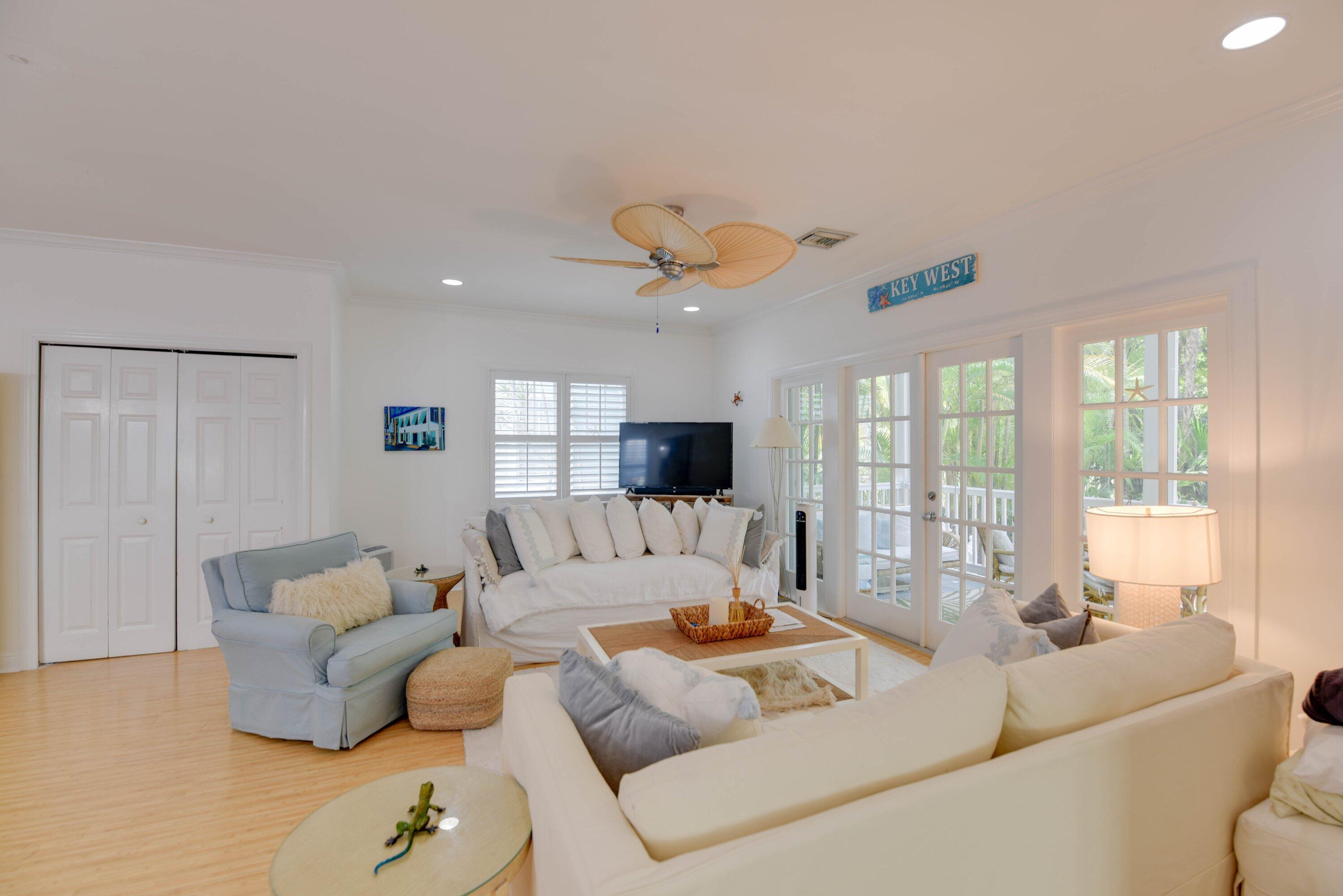Furnished living room with white sofas, a woven-blade ceiling fan, and French doors overlooking tropical greenery.