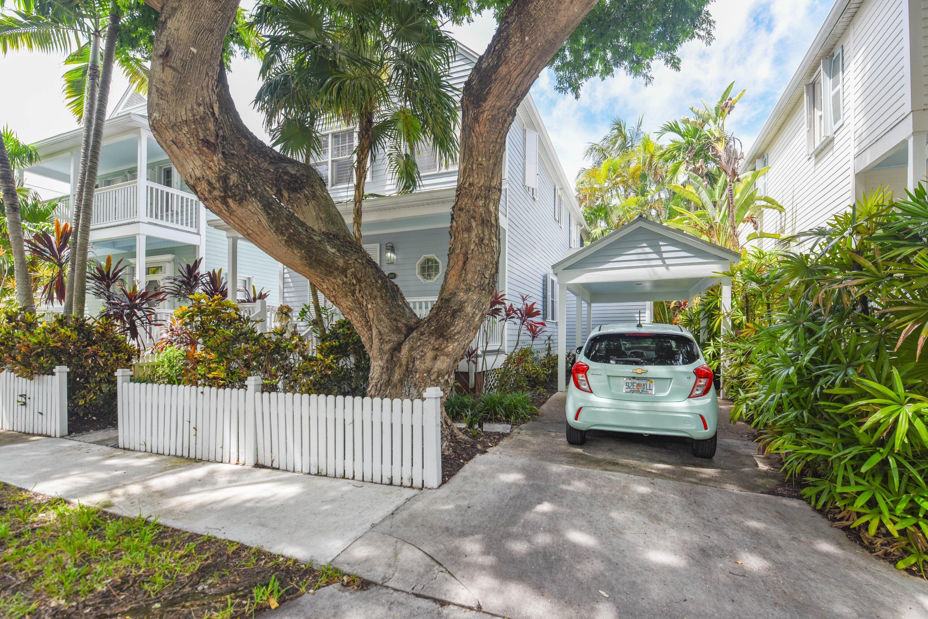 Light-blue home with white porches and a covered carport, framed by a large tree and tropical plants.