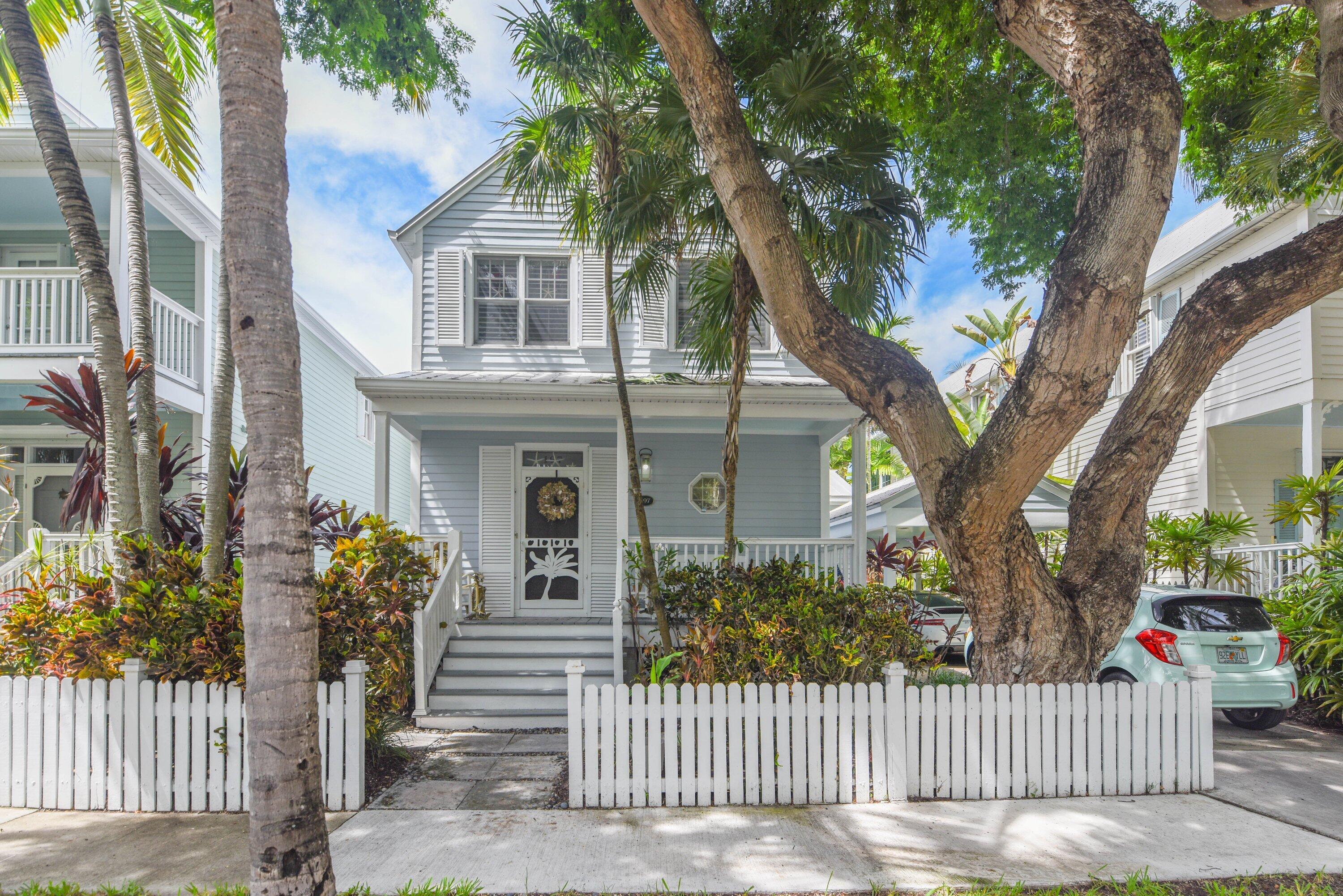 Two-story pale blue house behind a white picket fence, framed by palms and a large shade tree.