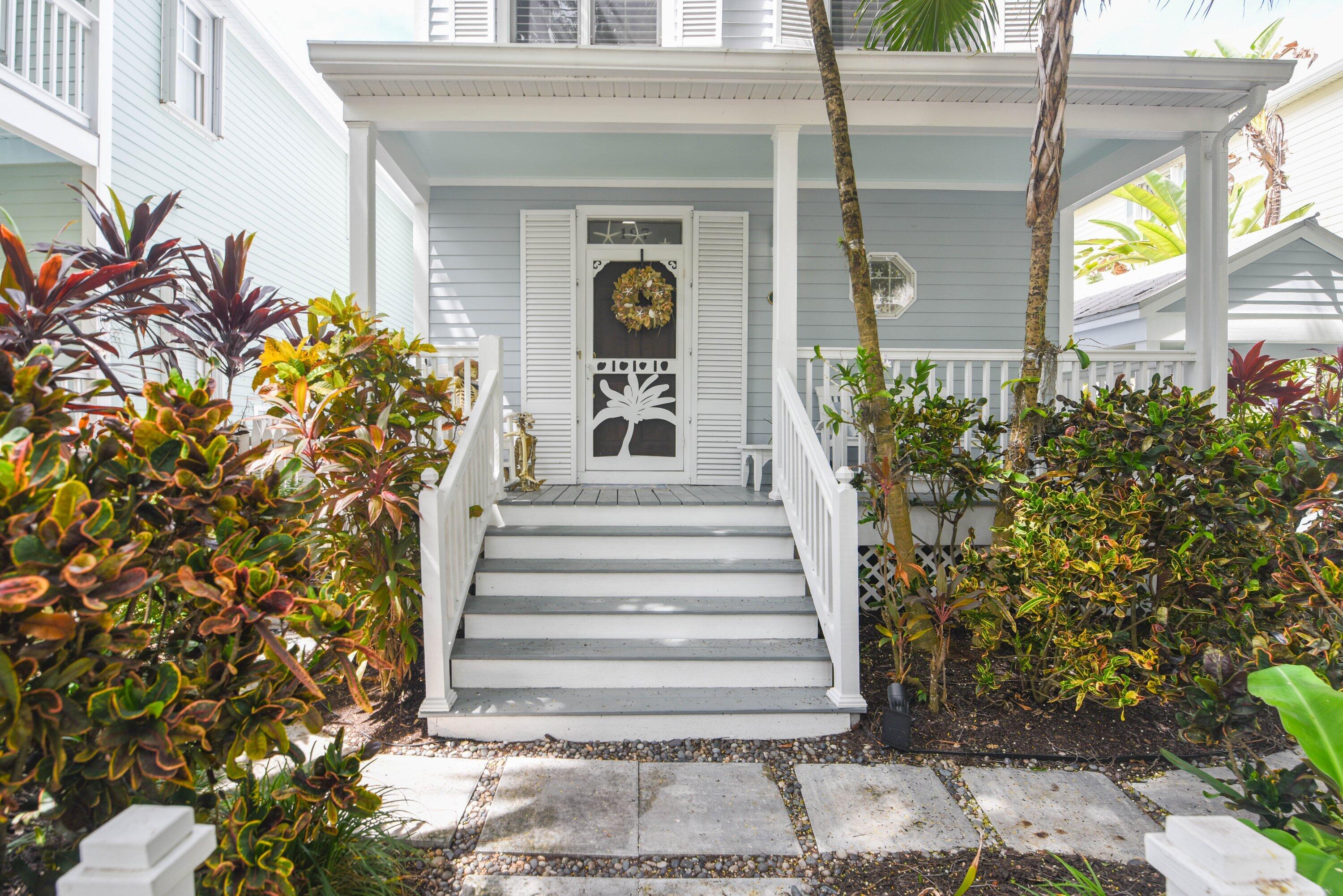 Light blue house entrance with white porch steps, shutters, and lush tropical plants.