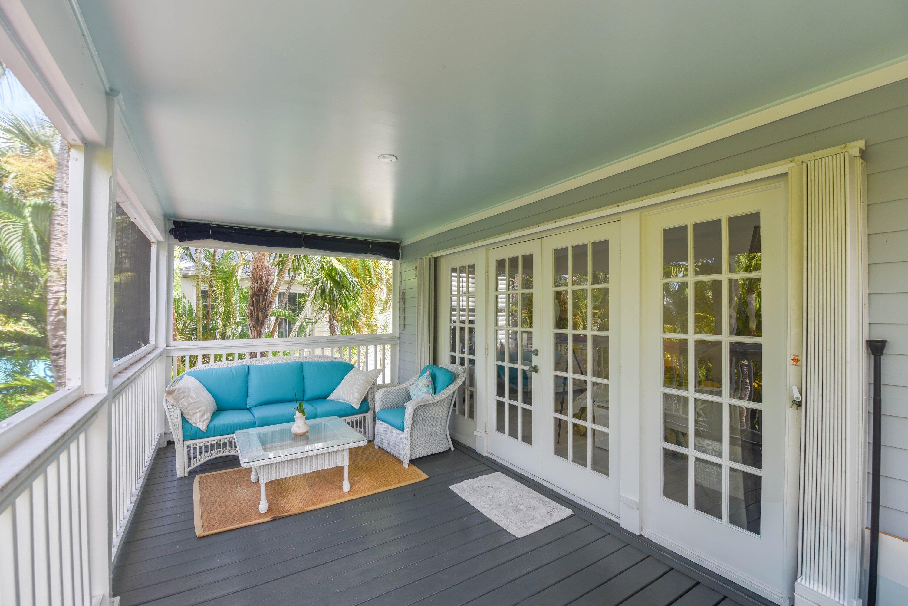 Screened porch with turquoise-cushioned wicker seating, white French doors, and palm trees beyond the railing.