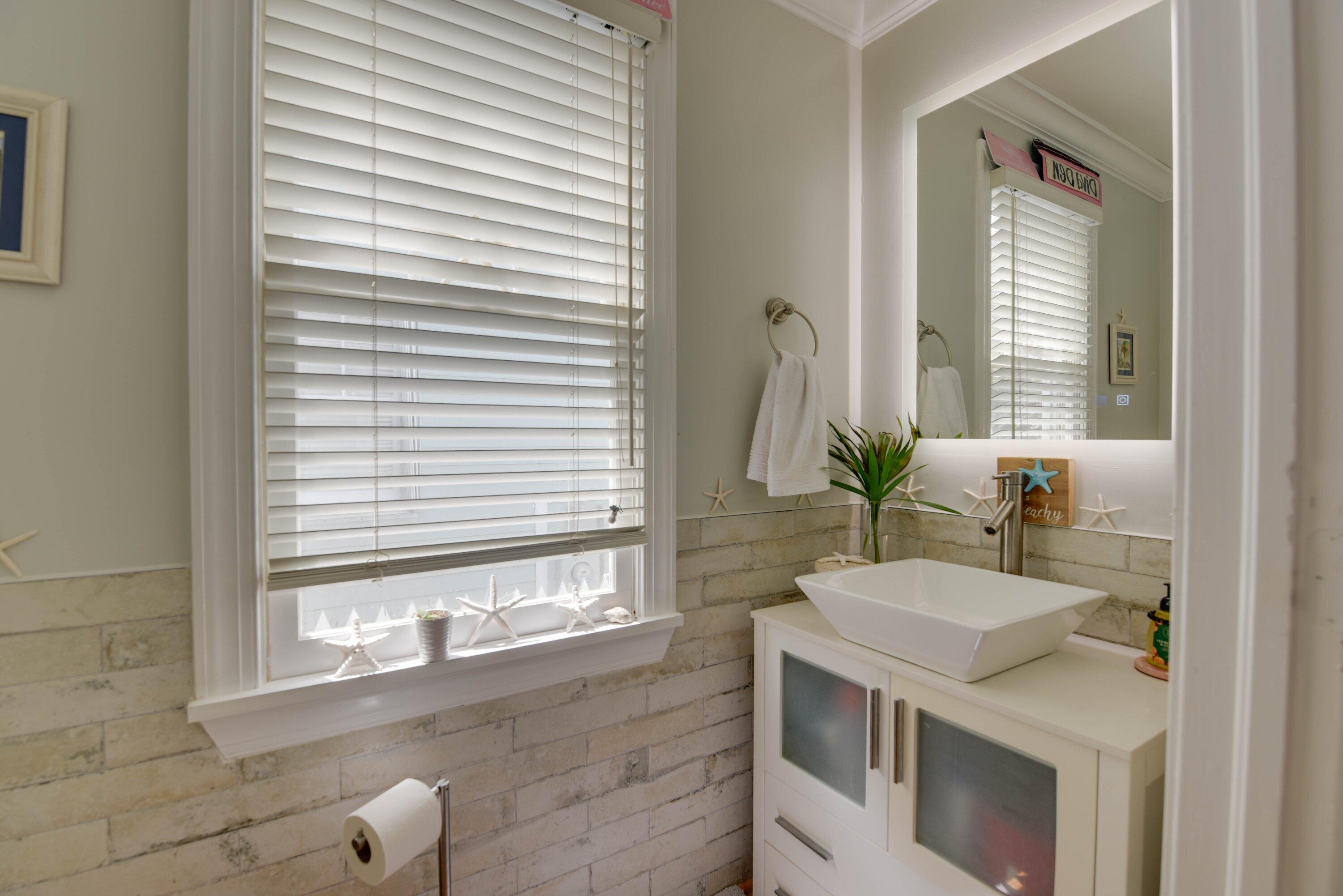 Bathroom vanity with a white vessel sink, illuminated mirror, pale stone tile, and coastal decorations.