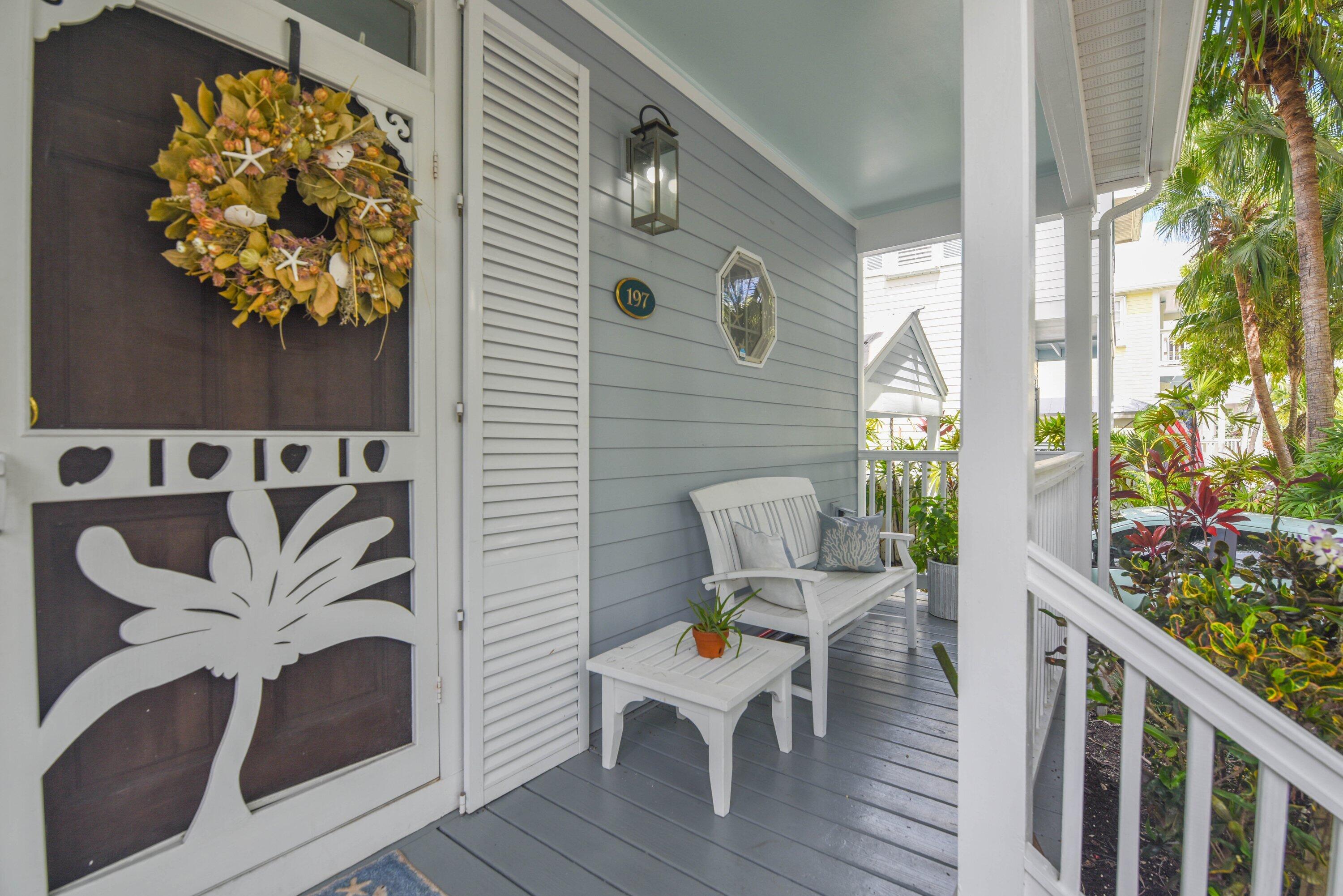 Covered porch with a white bench and table beside tropical landscaping.