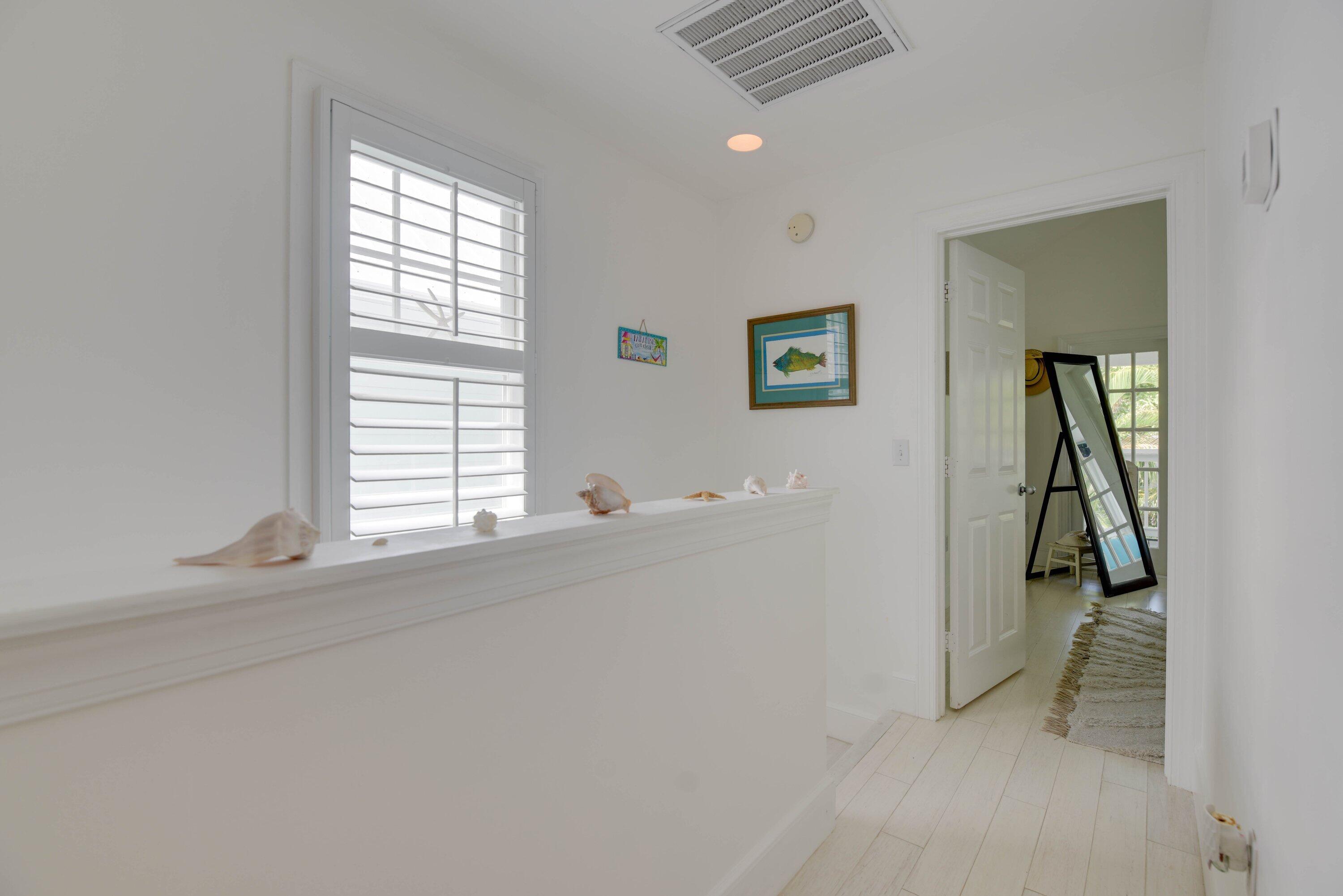 Bright white hallway with a shuttered window, seashells on the half wall, and a leaning mirror in the adjoining room.