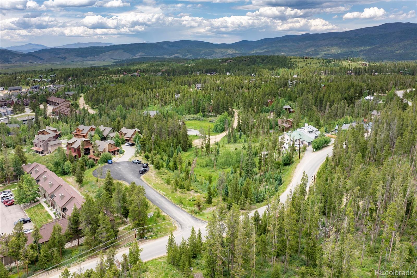 Aerial view of a wooded neighborhood, open lot, winding roads, and distant mountains.